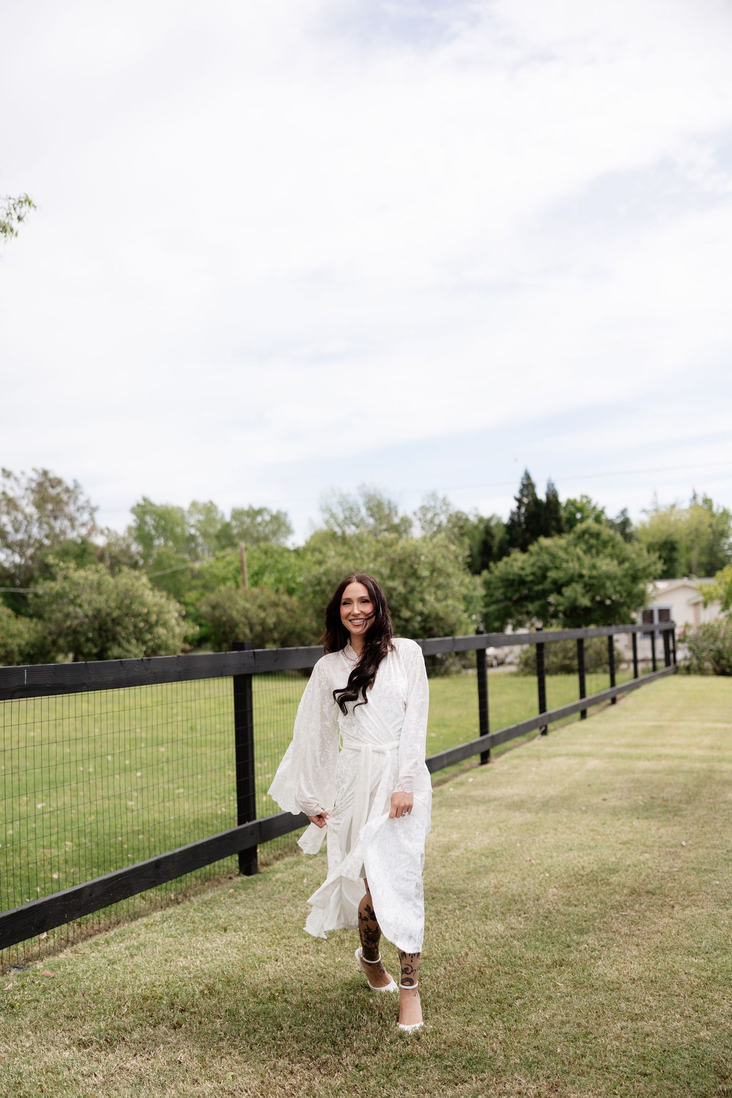 A smiling woman in a flowing white dress walks along a black fence on a green farm property.