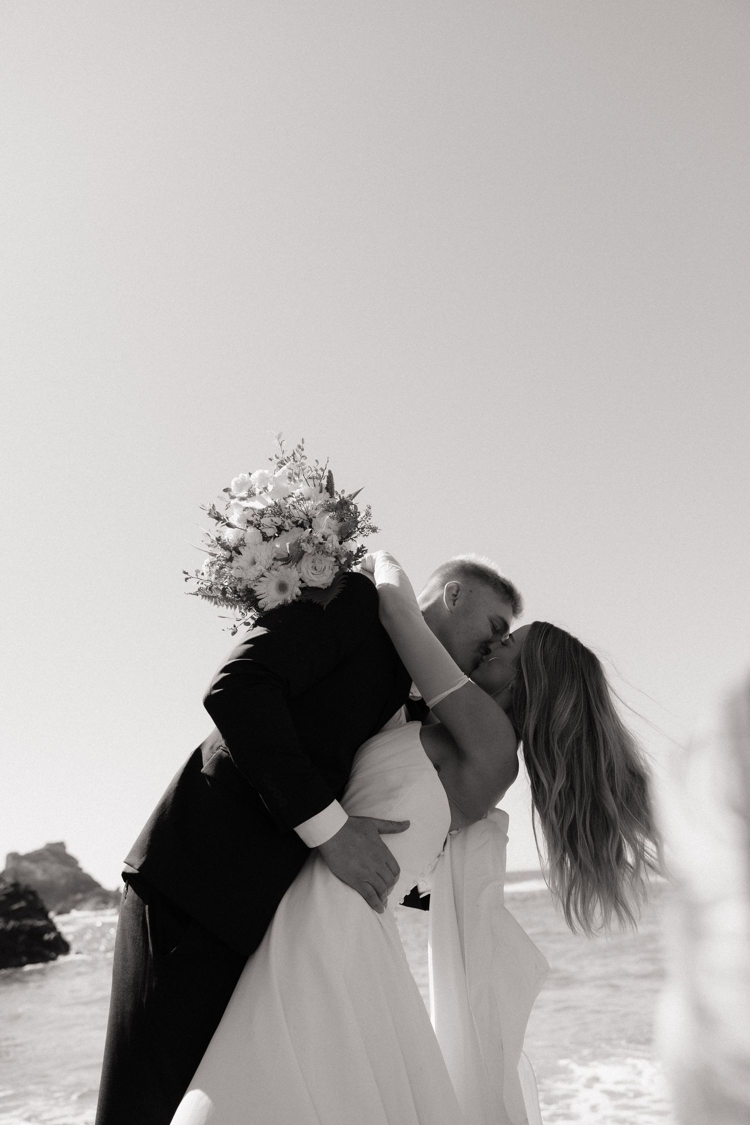 Black and white photo of a bride and groom kissing on a beach, bride holding a floral bouquet, ocean in background.