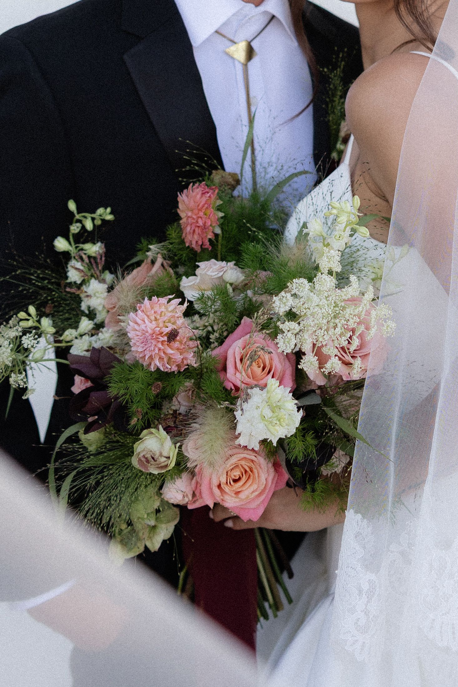 Bride and groom holding a lush pink and white wedding bouquet with roses and dahlias, bride wearing lace dress with veil.