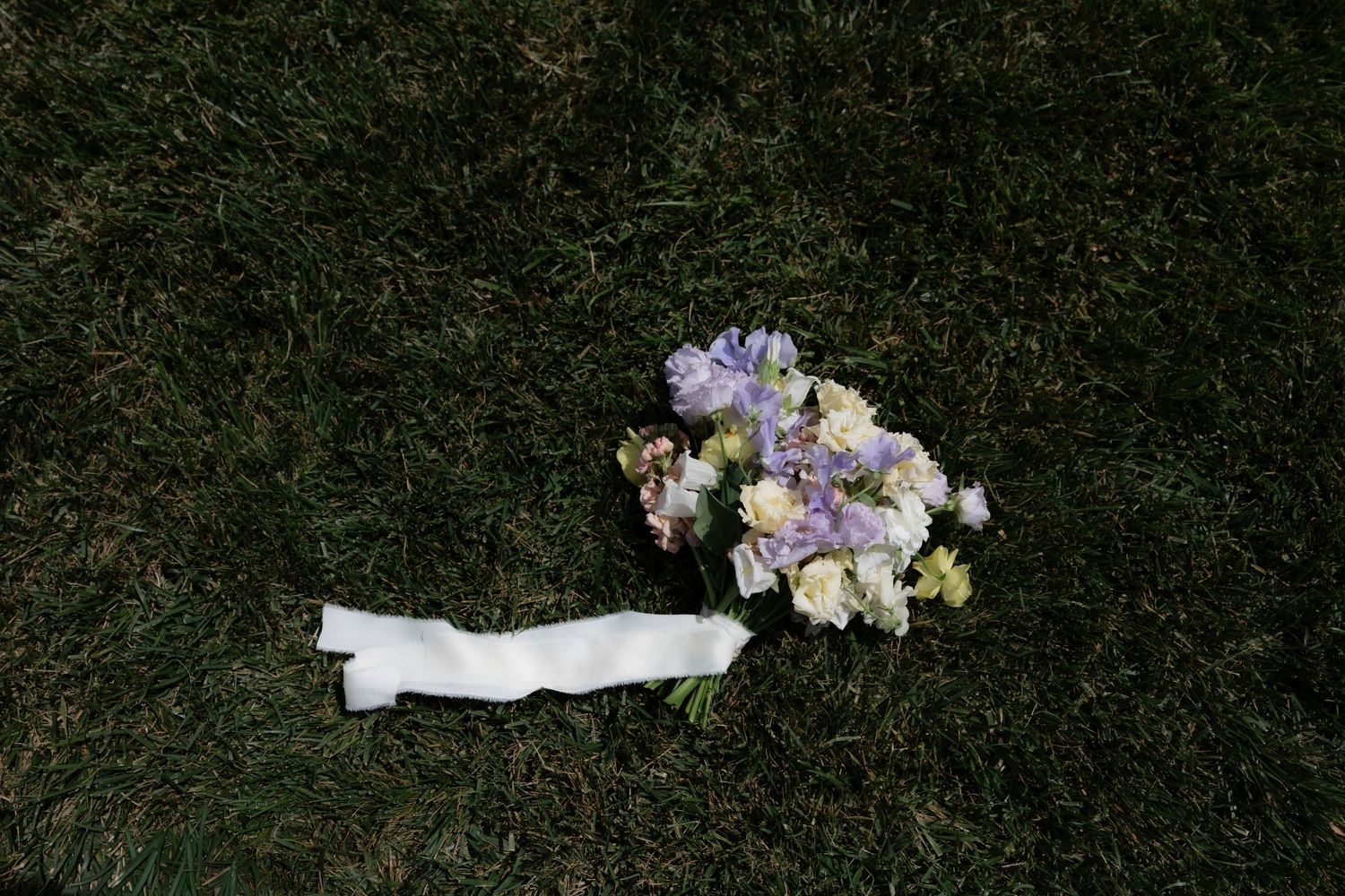 Delicate bridal bouquet of lavender and white flowers with white ribbon lying on lush green grass.