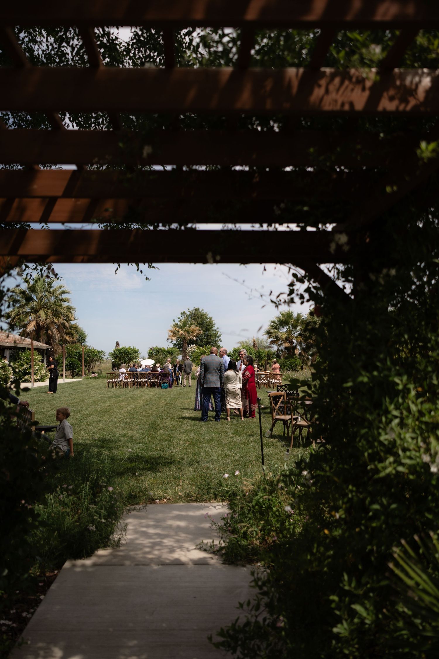 Outdoor wedding venue viewed through wooden pergola with guests mingling on green lawn under blue sky.