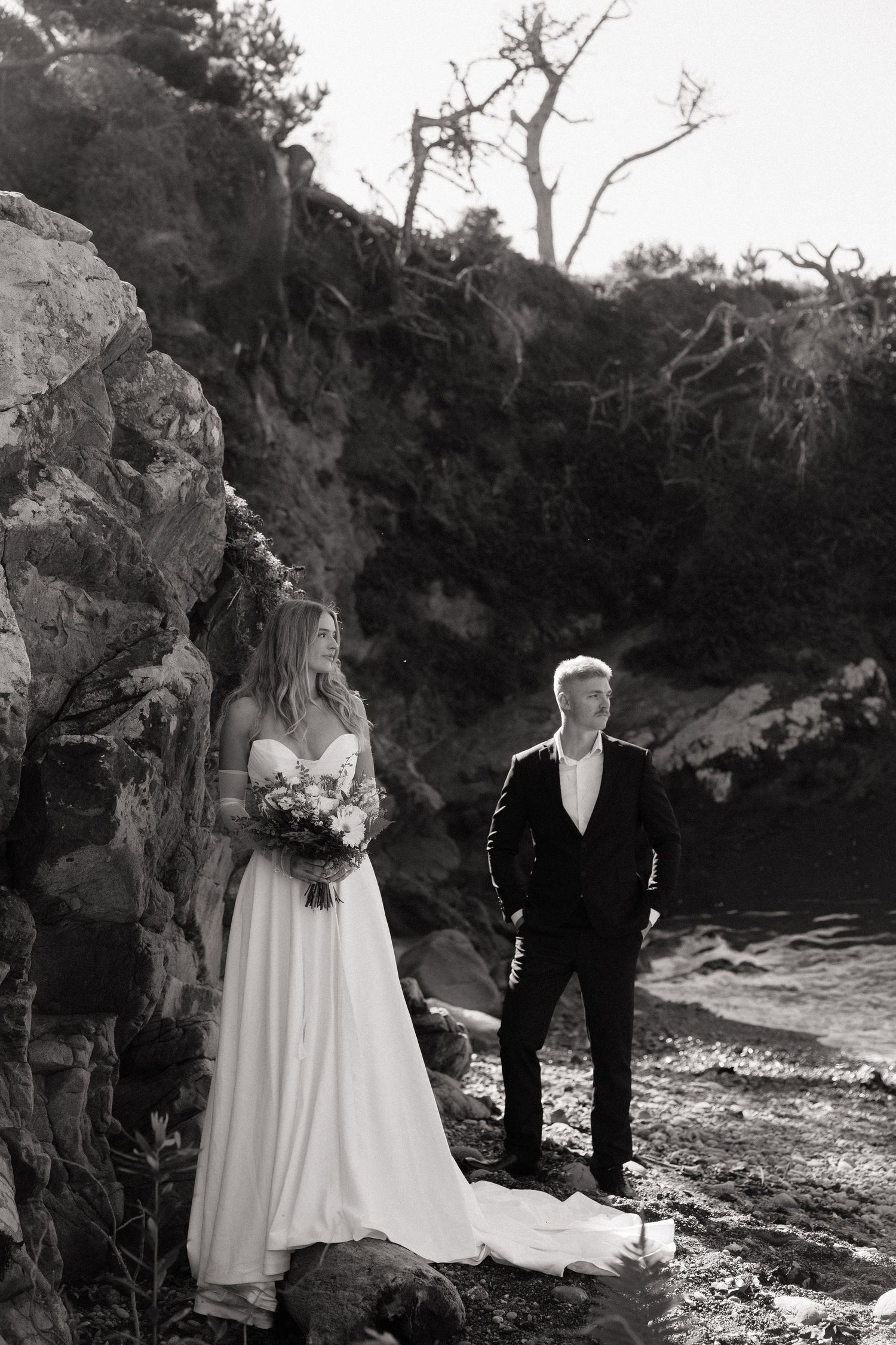 Black and white photo of bride and groom standing by rocky coastal cliffs, bride holding bouquet in elegant gown.