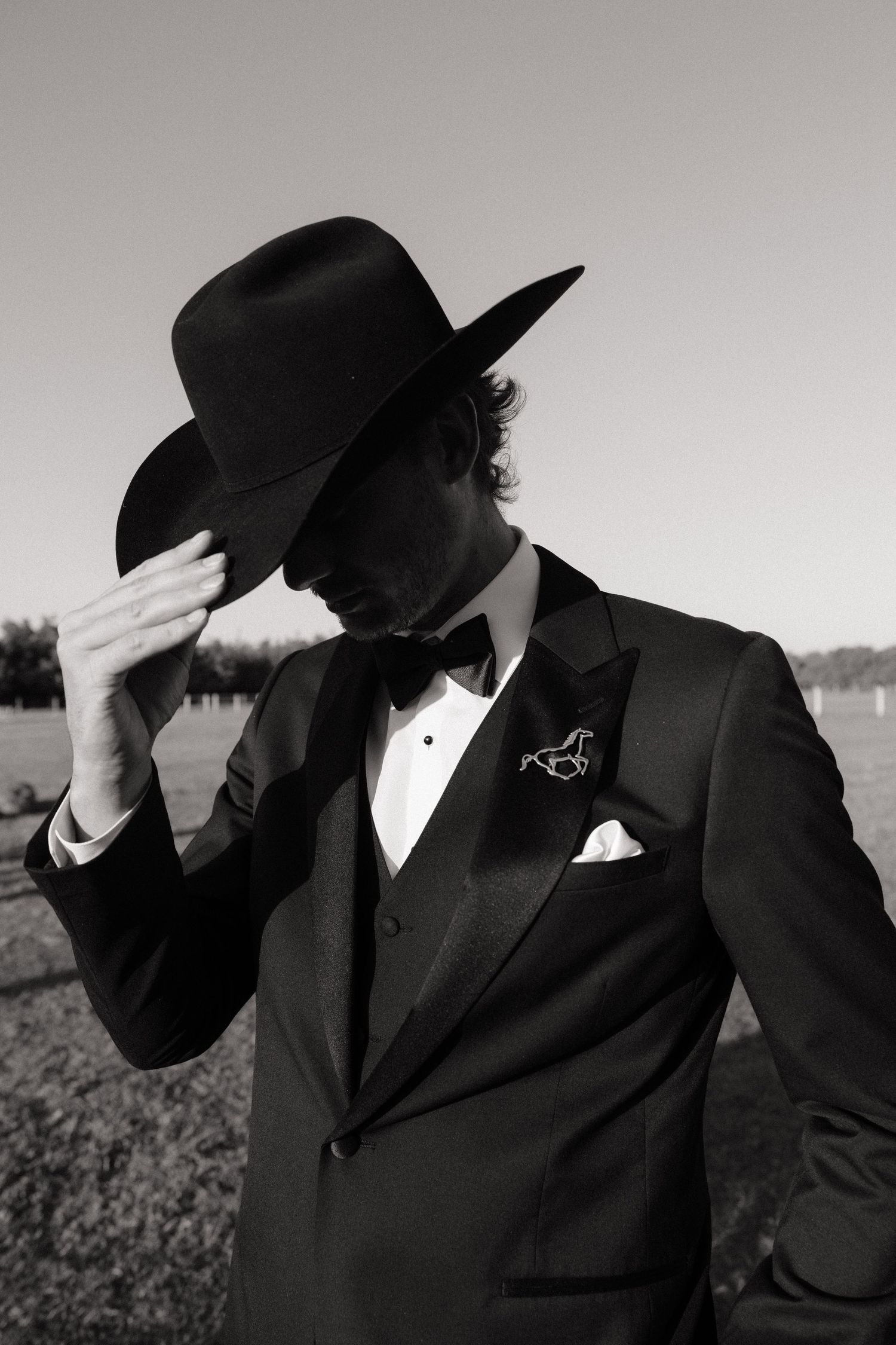 Black and white photo of a man in a tuxedo and cowboy hat, adjusting his hat outdoors.