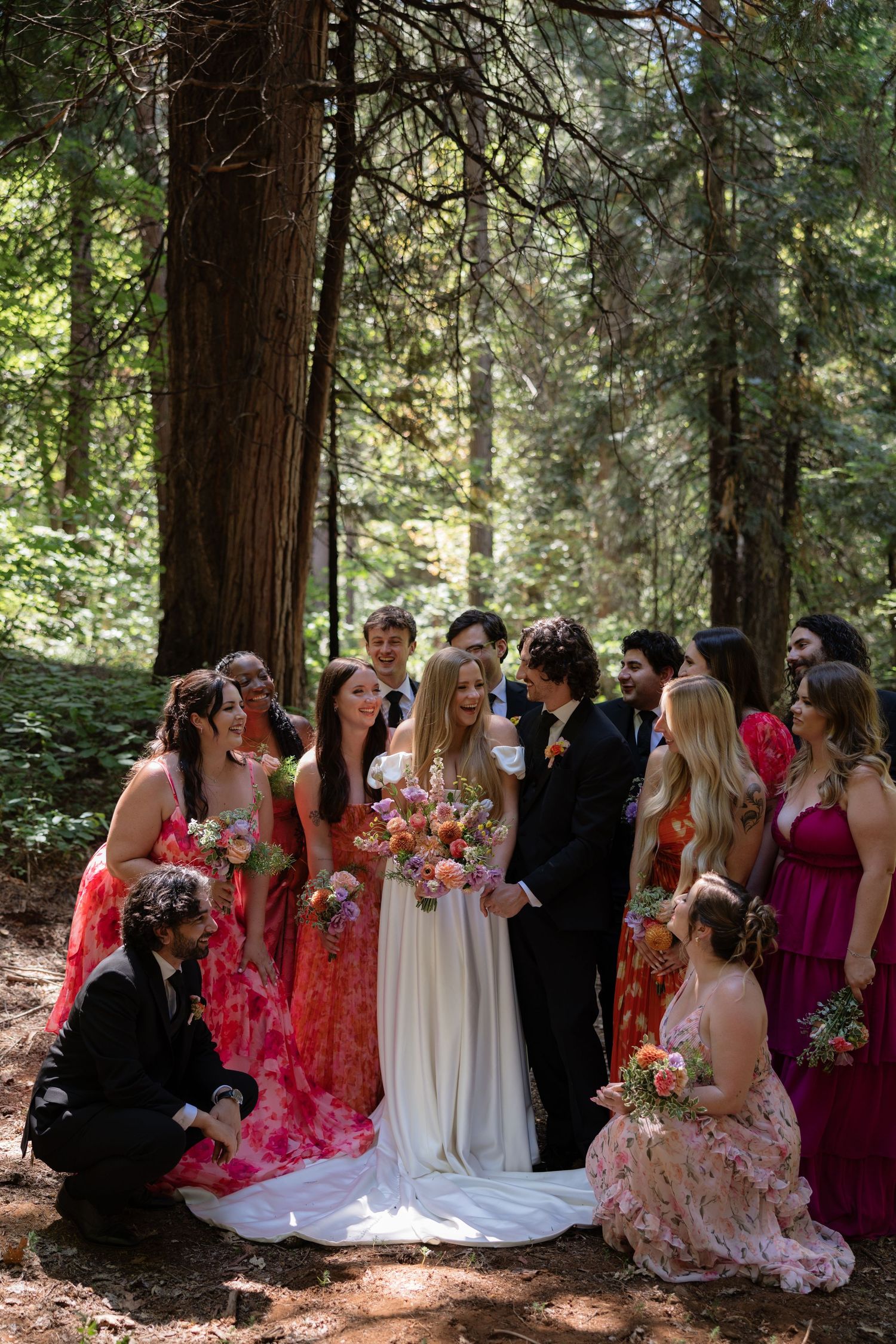Wedding party in colorful pink dresses gathered around bride and groom in a sunlit forest setting.