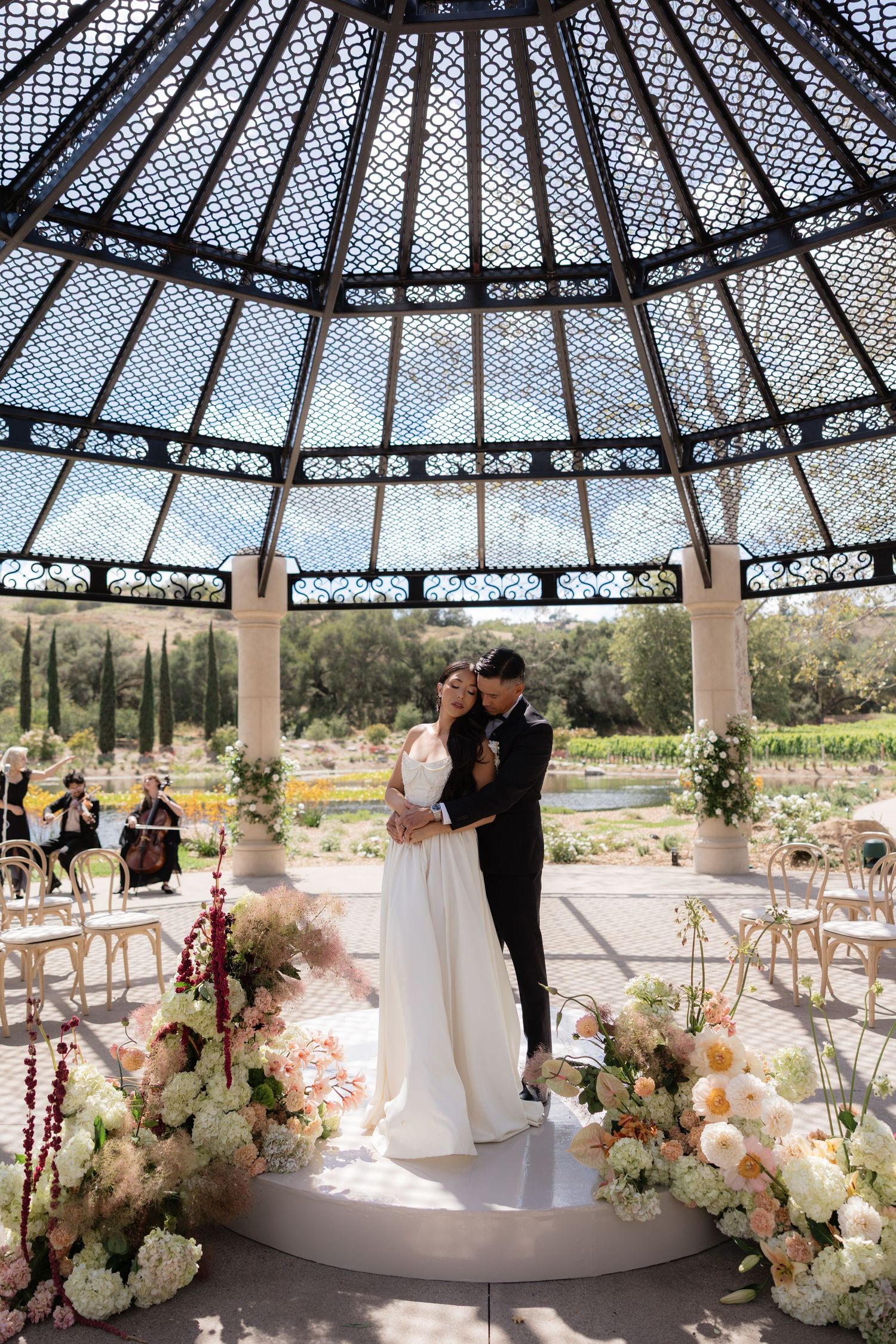Bride and groom embracing under a stunning glass dome gazebo surrounded by lush floral arrangements.