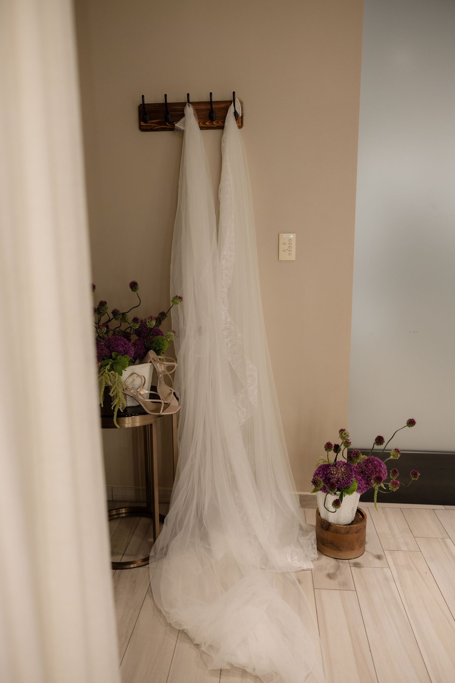 A long white wedding veil and dress hang on a wooden wall hook, surrounded by purple floral arrangements on a light wood floor.