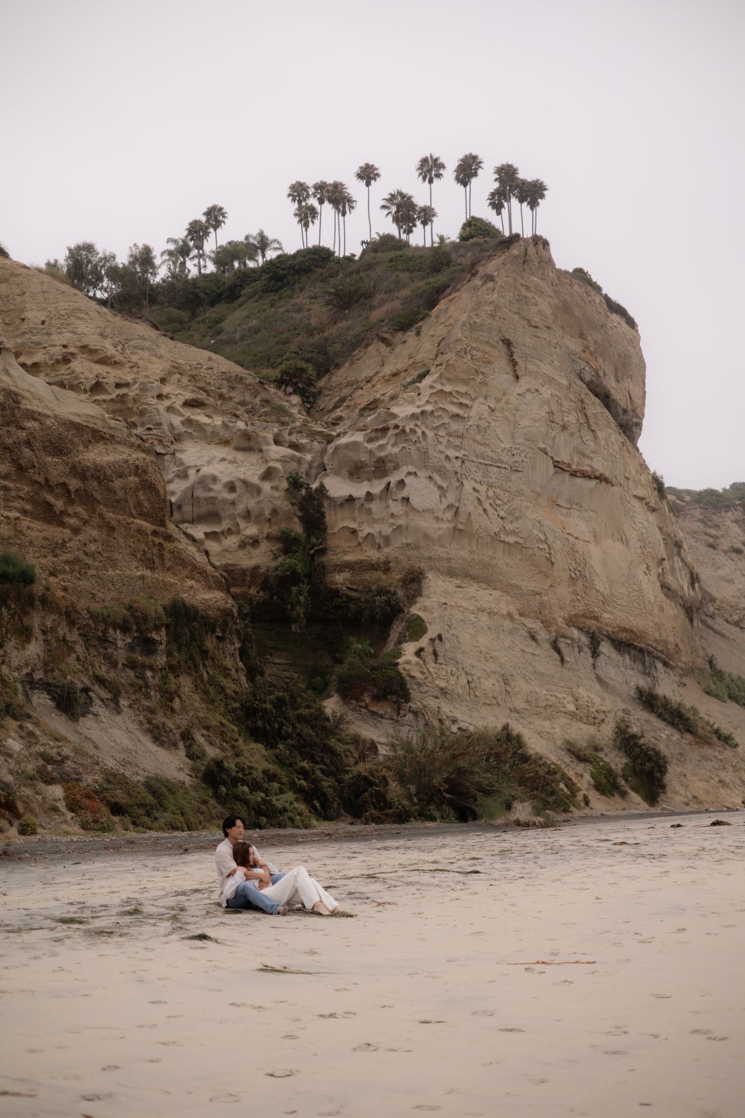 Couple sitting together on a sandy beach below towering sandstone cliffs topped with palm trees.