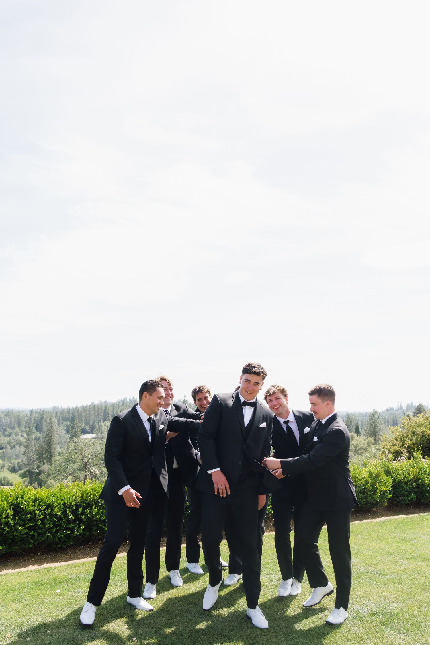 Groom and groomsmen in black tuxedos with white sneakers gathered on green lawn.