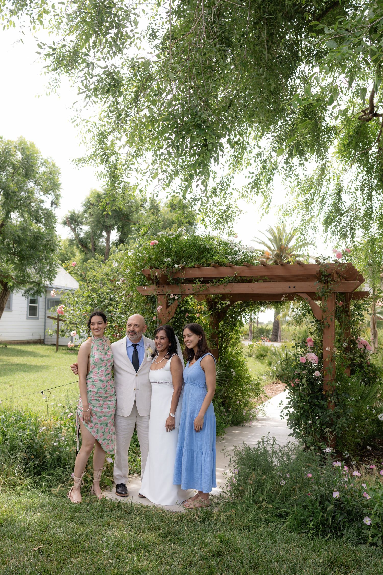 Wedding party of four posing under lush pergola with climbing roses in a beautiful garden setting.