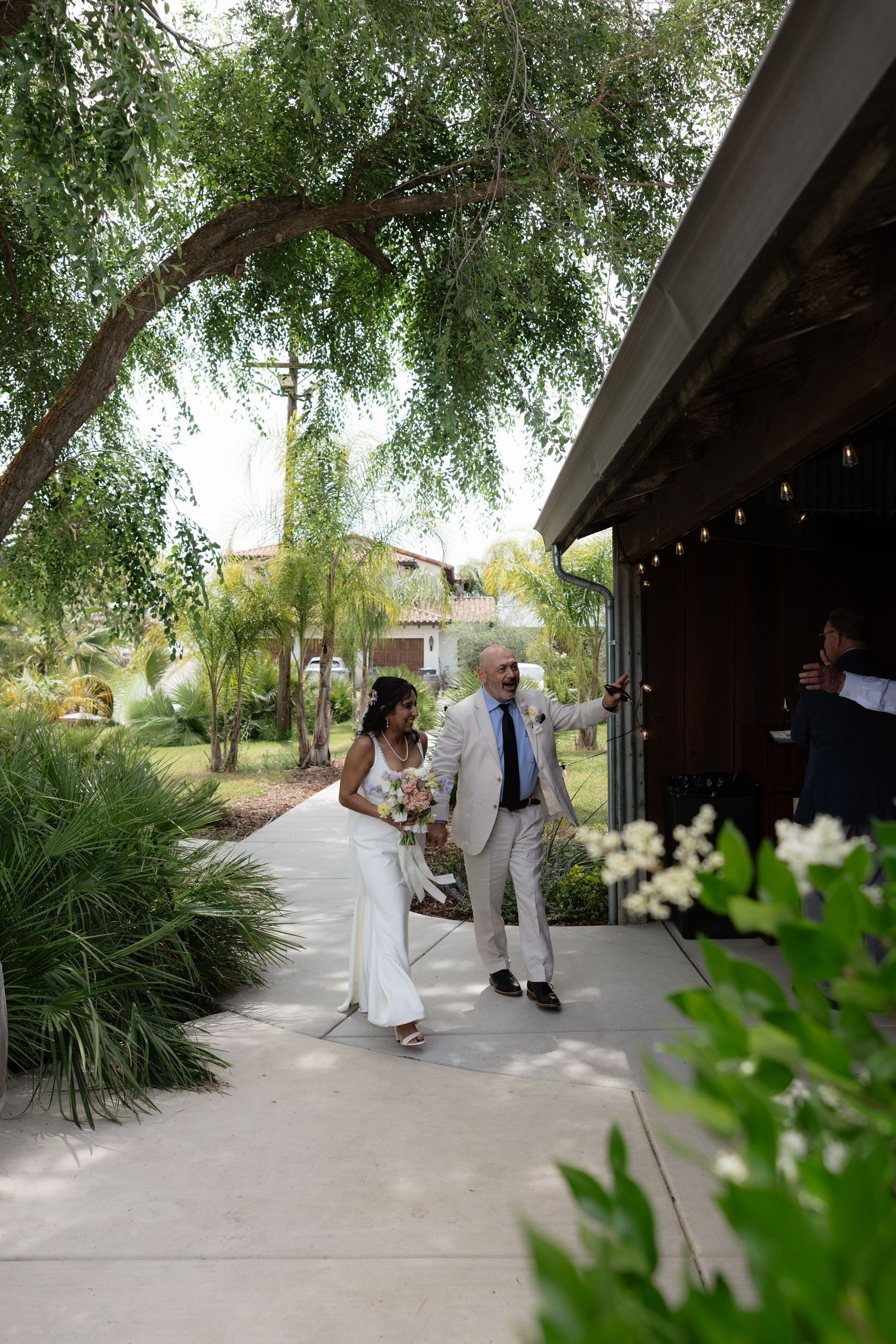 Bride and groom walk together along a garden path lined with tropical plants toward a warmly lit wedding reception venue.