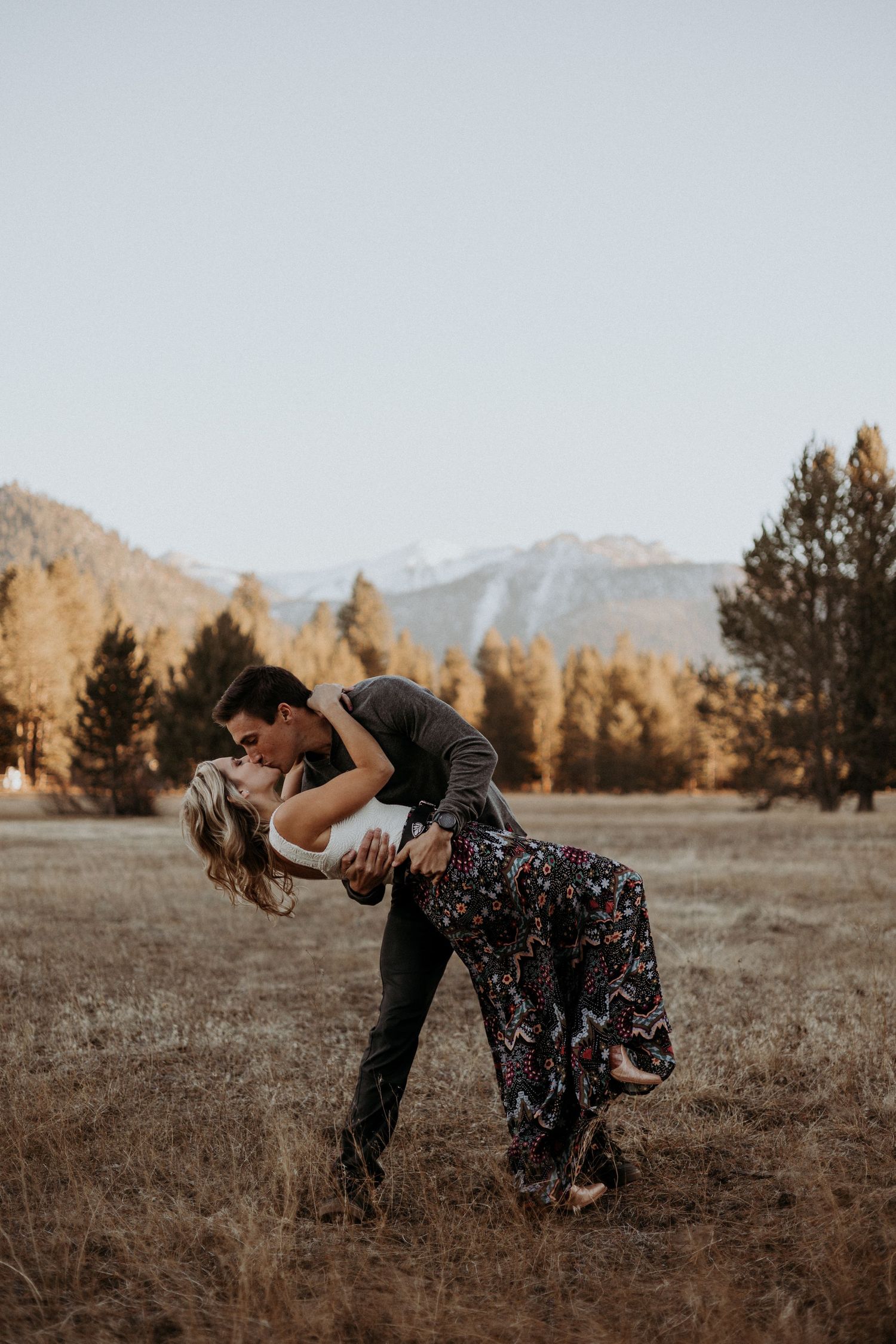 A couple shares a passionate dip kiss in a golden autumn field with snow-capped mountains in the background.