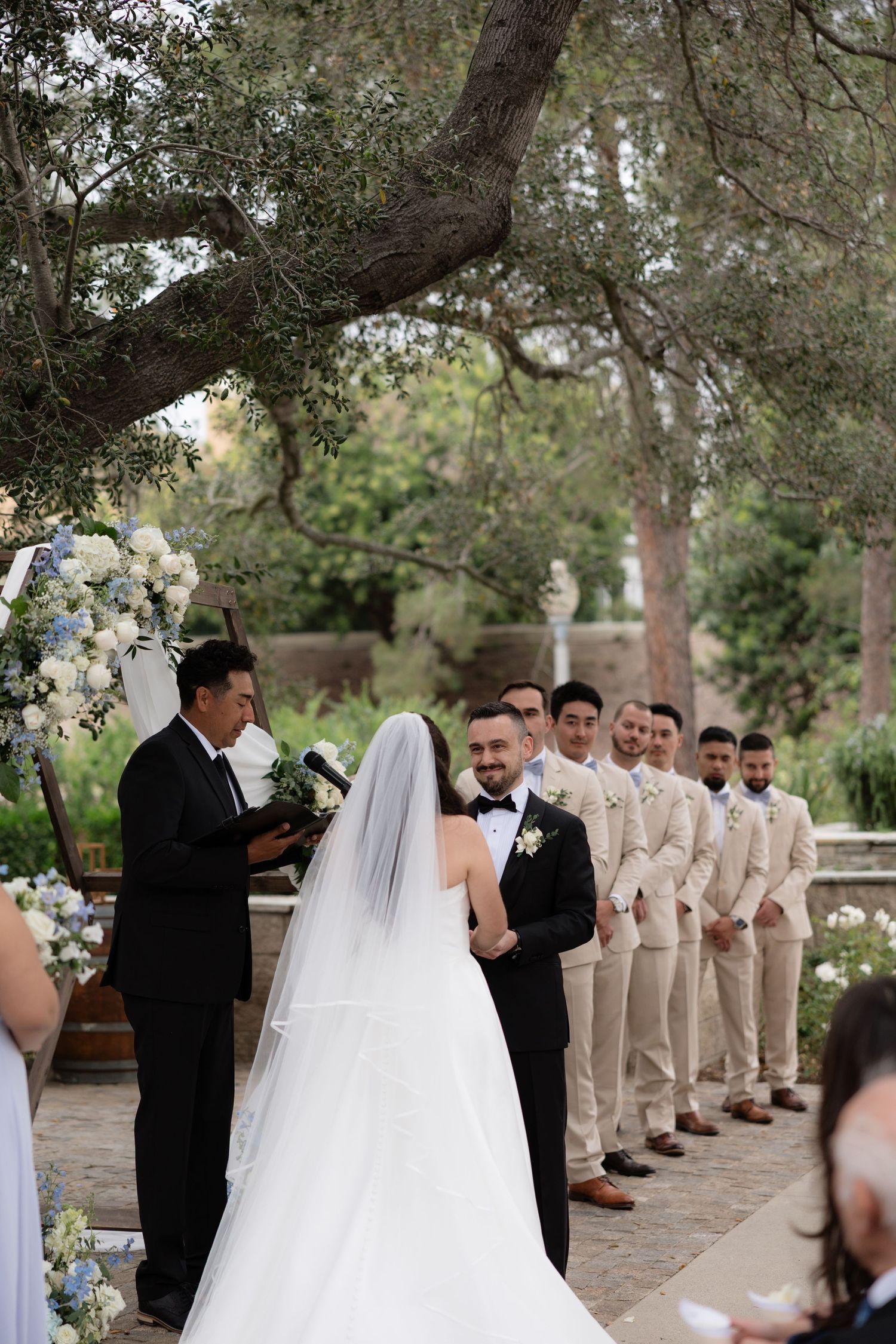 Bride and groom exchanging vows under an olive tree with groomsmen in tan suits during outdoor ceremony.