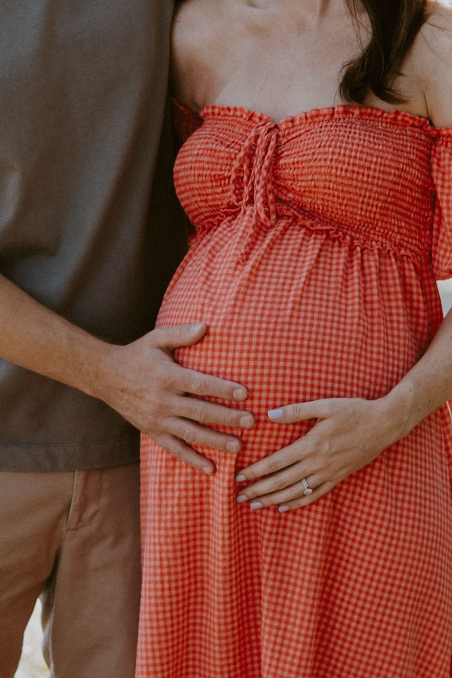 Couple cradling pregnant belly, woman wearing red gingham off-shoulder maternity dress outdoors.