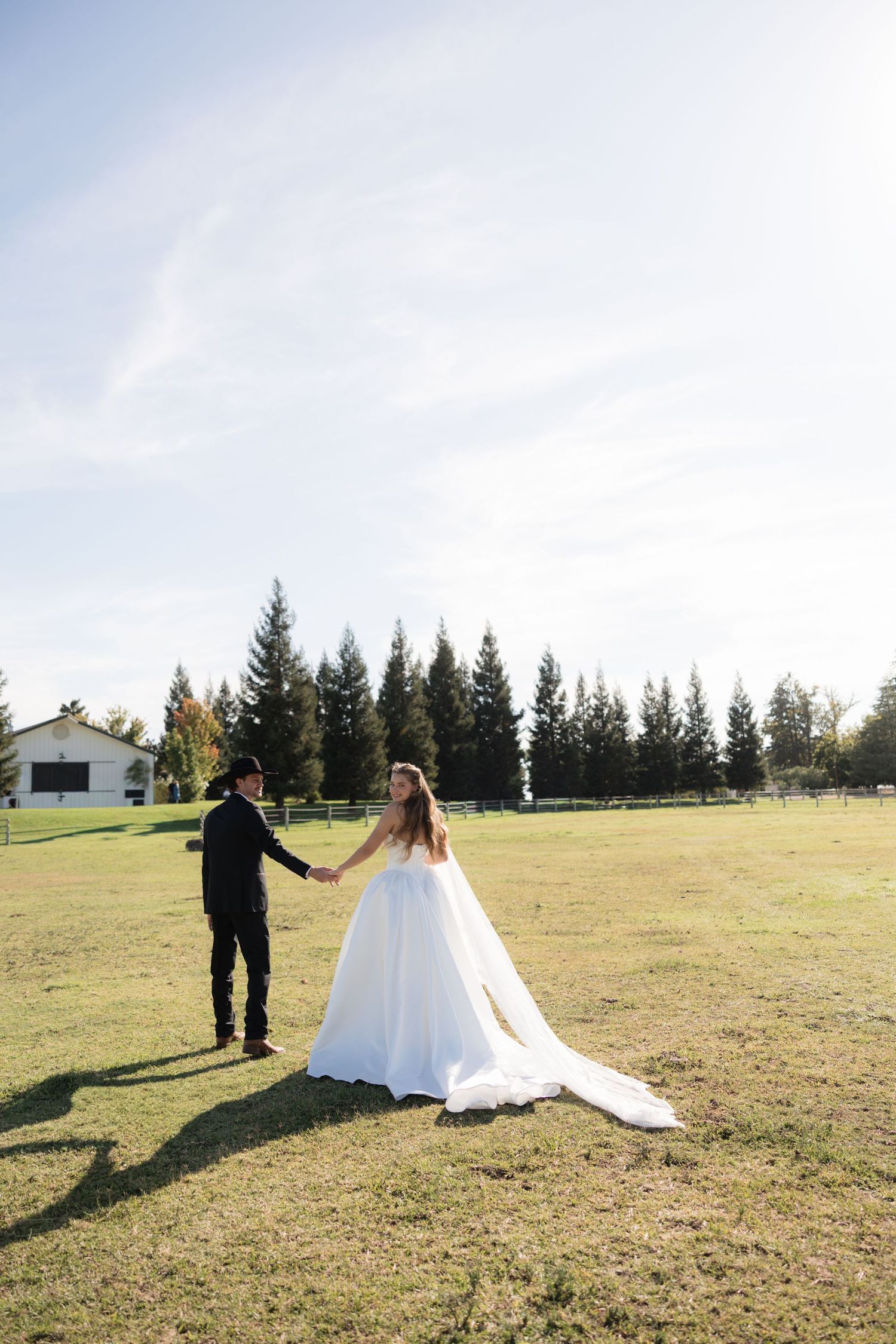 Bride in white gown and groom in black suit hold hands in a sunlit green field with tall pine trees and a barn.