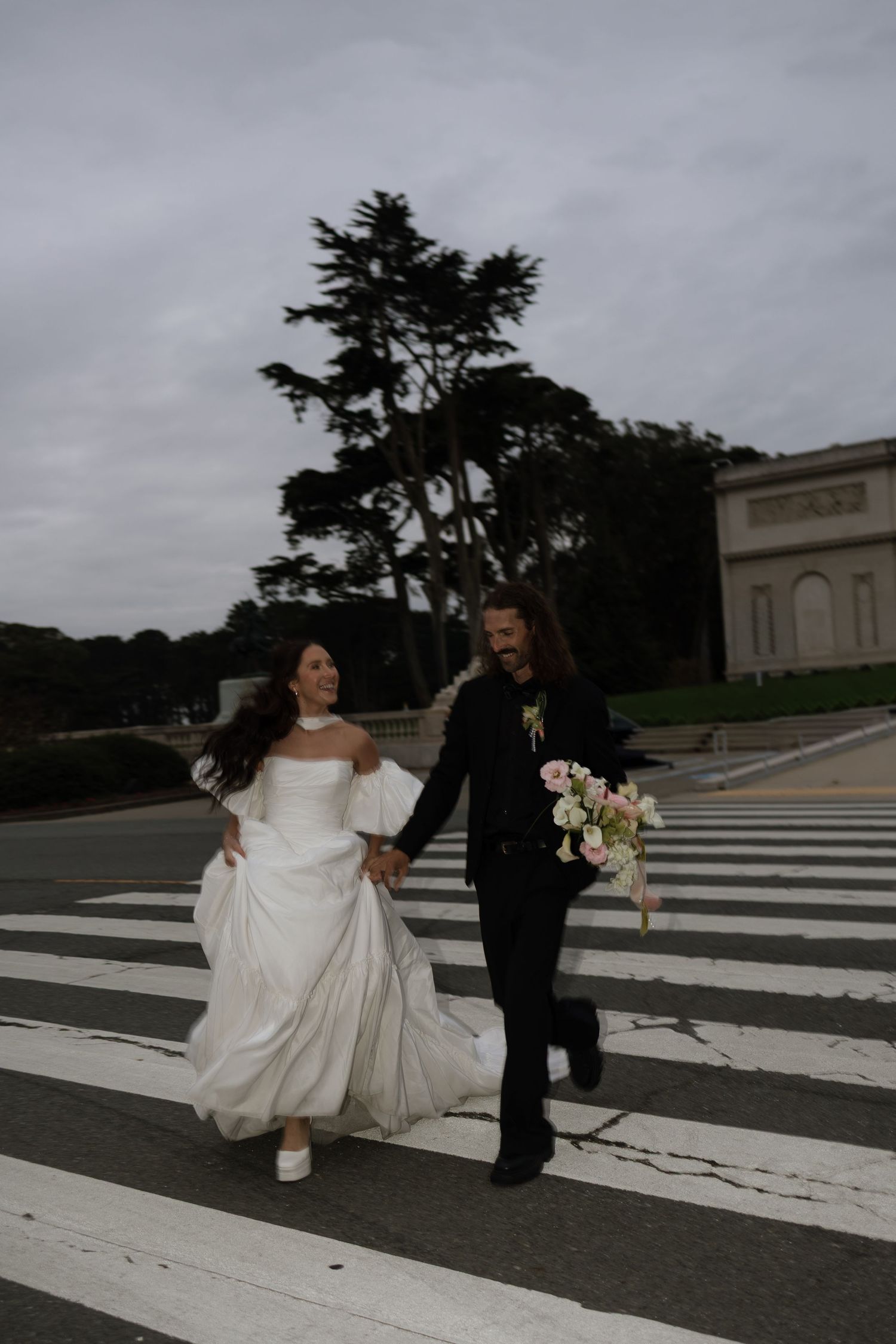 Bride and groom joyfully crossing street holding hands near neoclassical building on cloudy wedding day.