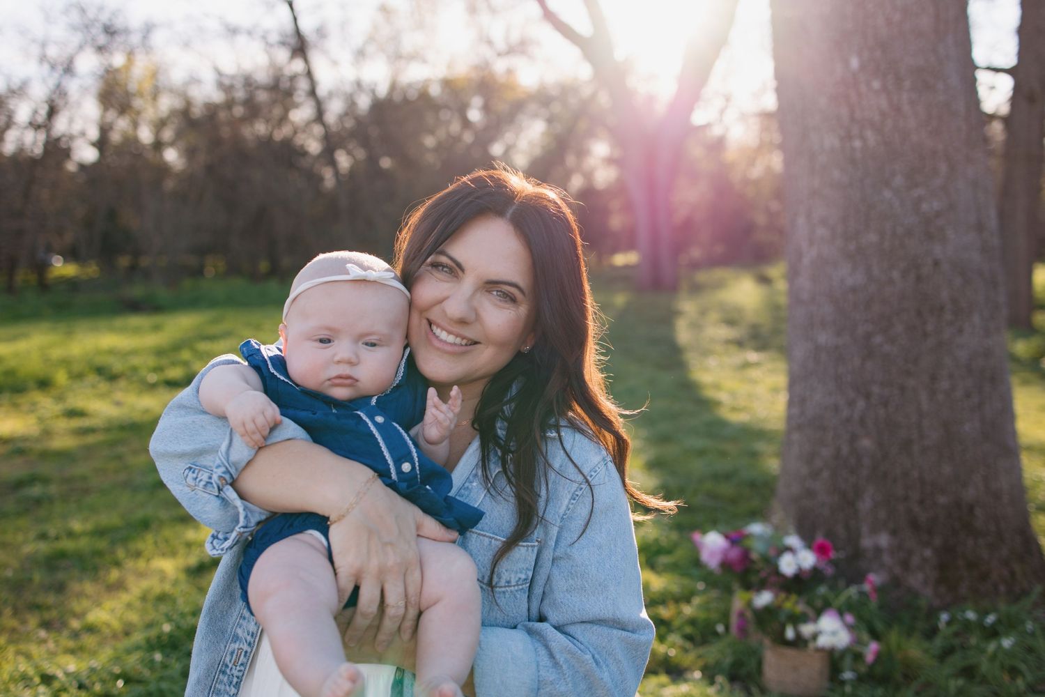 Smiling brunette mother holding infant baby outdoors in golden sunlight among green trees and flowers.