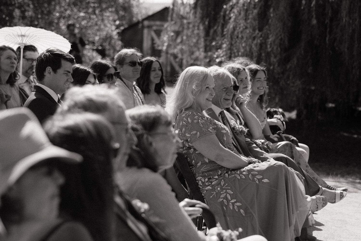 Black and white photo of wedding guests seated outdoors, enjoying a sunlit garden ceremony.
