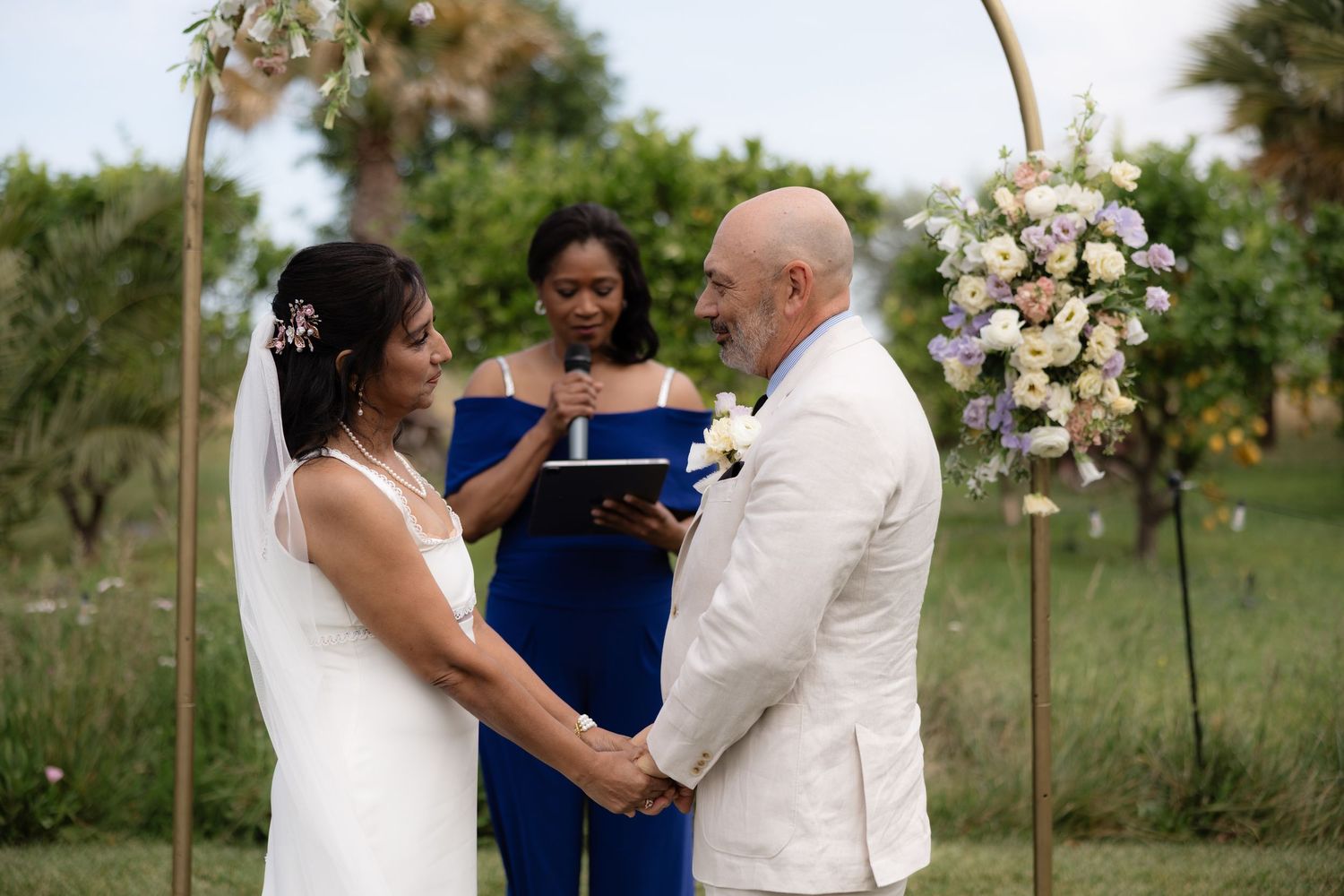 Bride and groom exchange vows at outdoor wedding ceremony with circular floral arch and officiant.