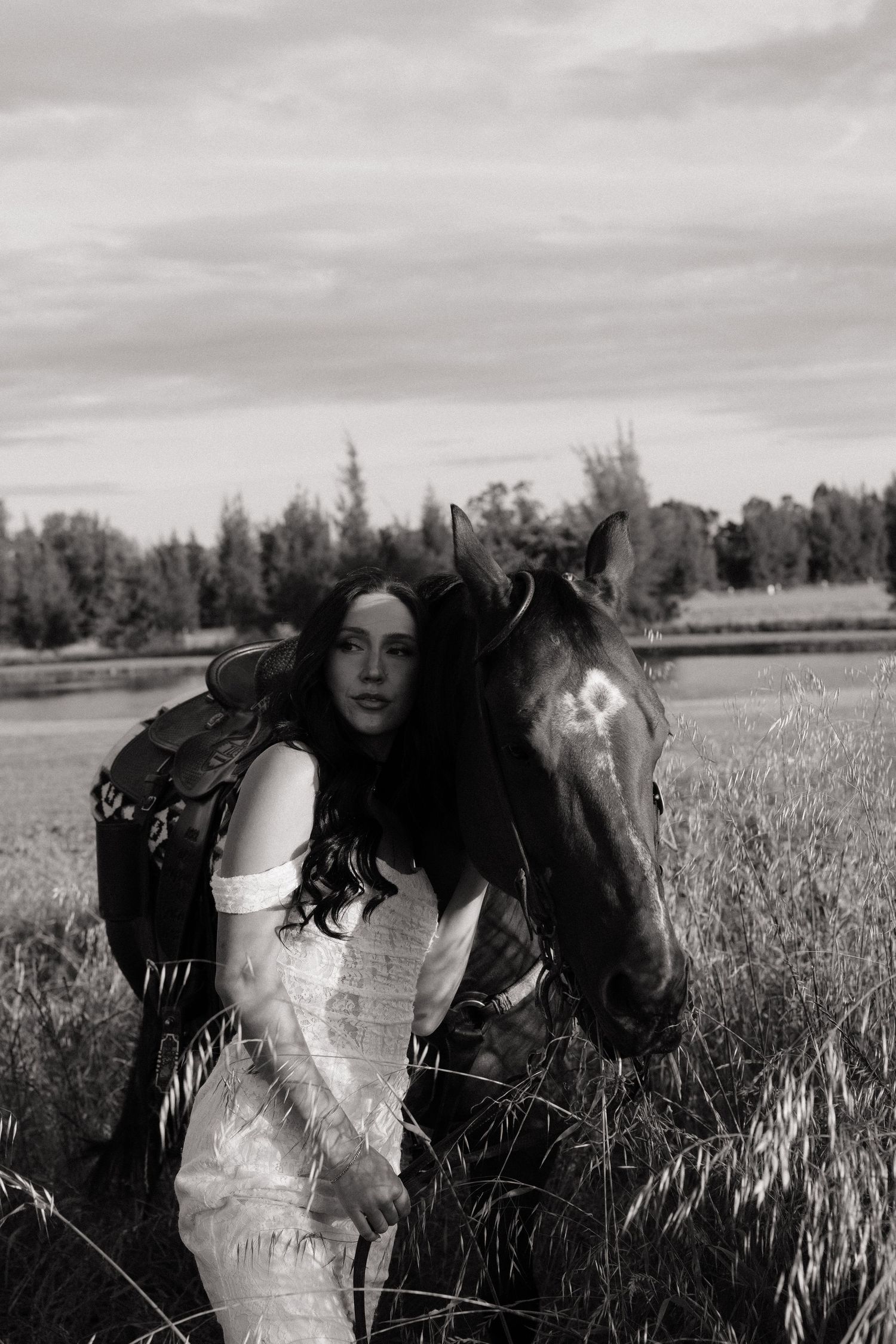 Bride in white lace gown standing beside a dark horse in a scenic open field.