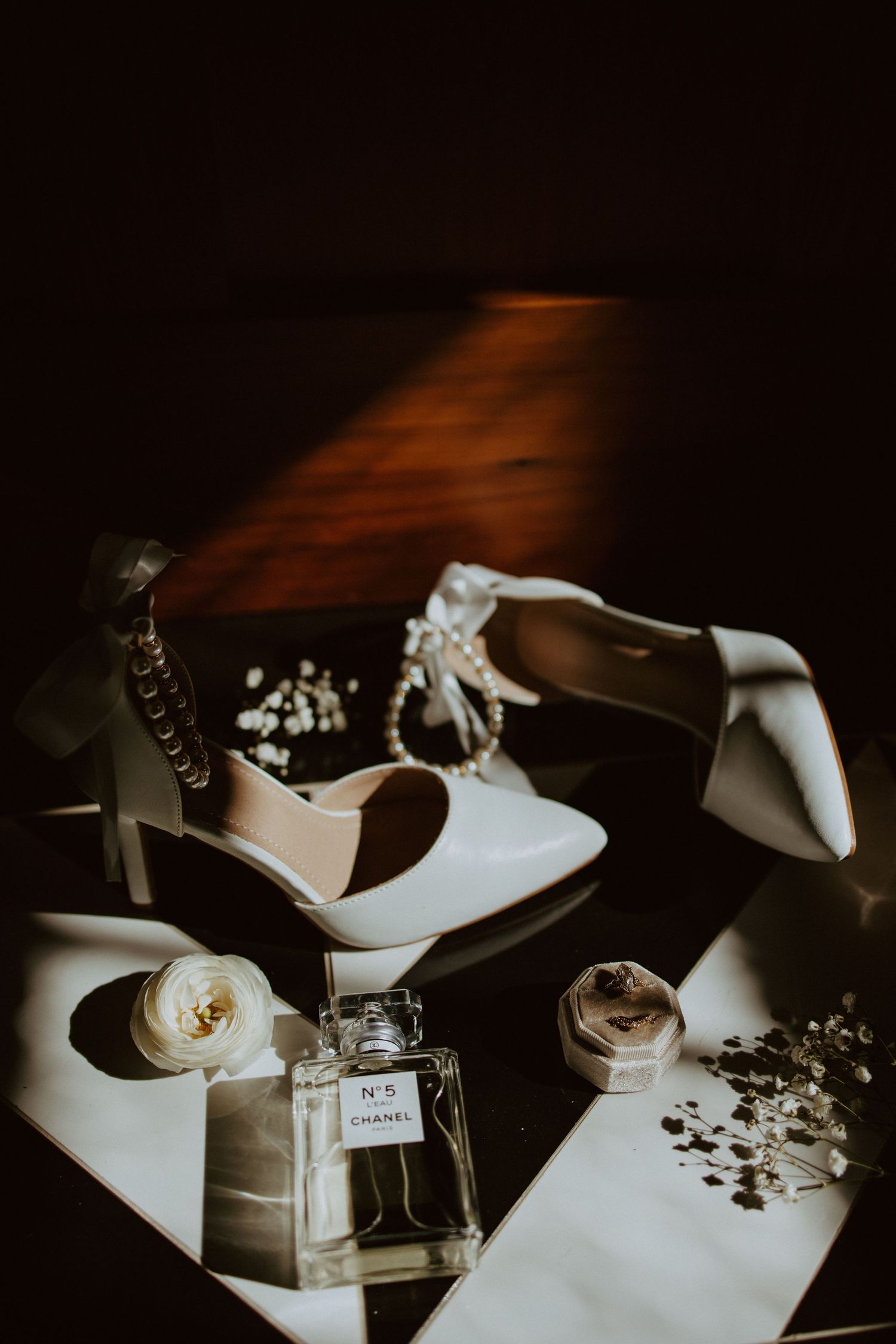 White bridal heels, Chanel No.5 perfume, pearl bracelet, and floral ring box in moody lighting.