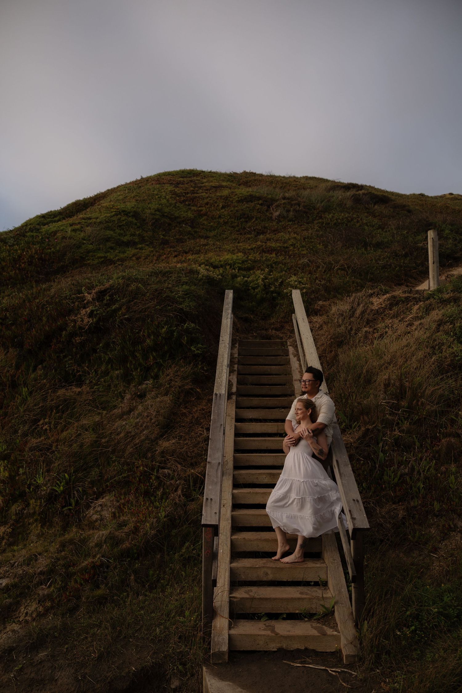 Couple embracing on wooden beach stairs, green grassy hillside, overcast sky, woman in white dress, romantic outdoor portrait.