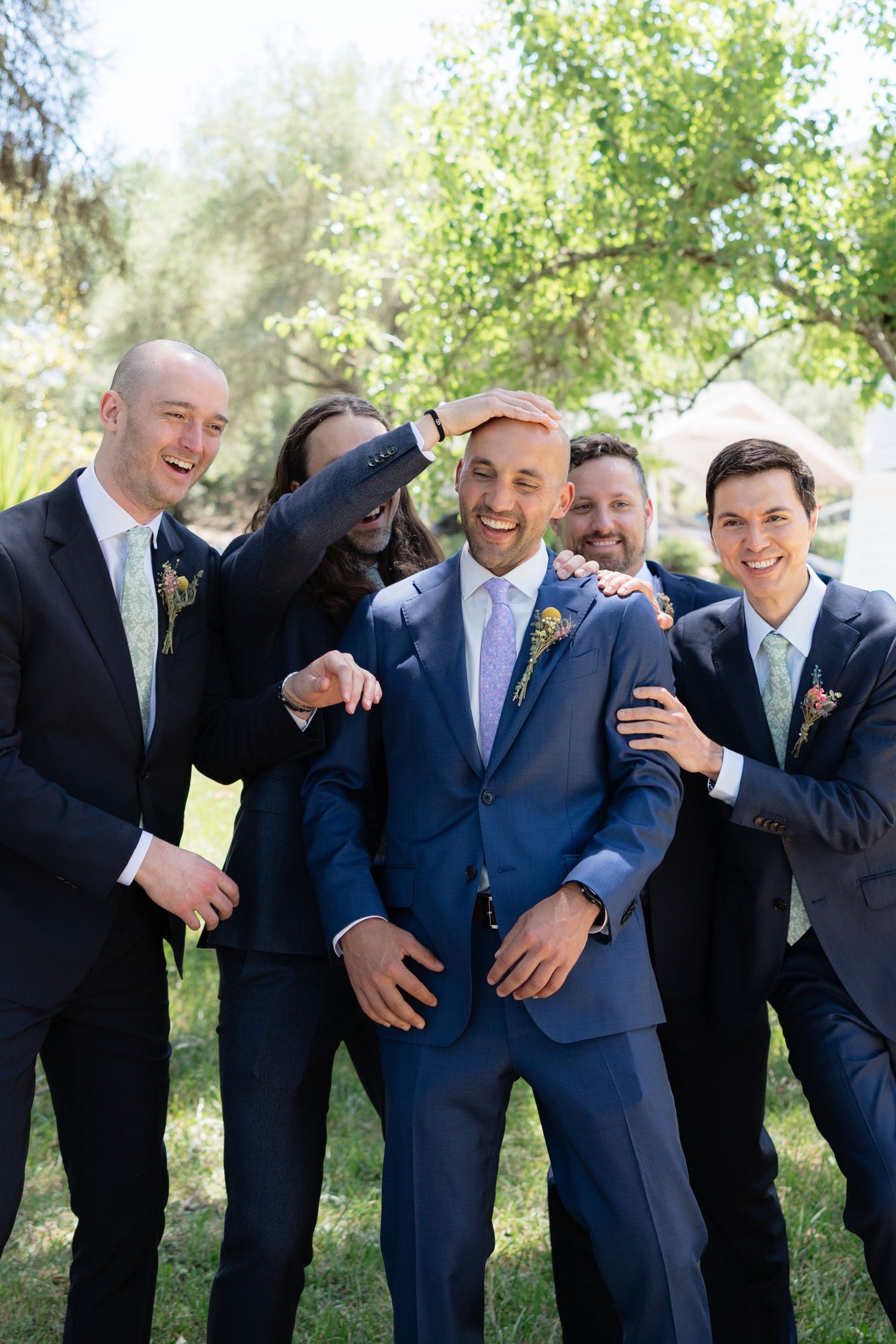 Groom laughing with groomsmen in navy suits playfully posing together outdoors in a sunny garden setting.