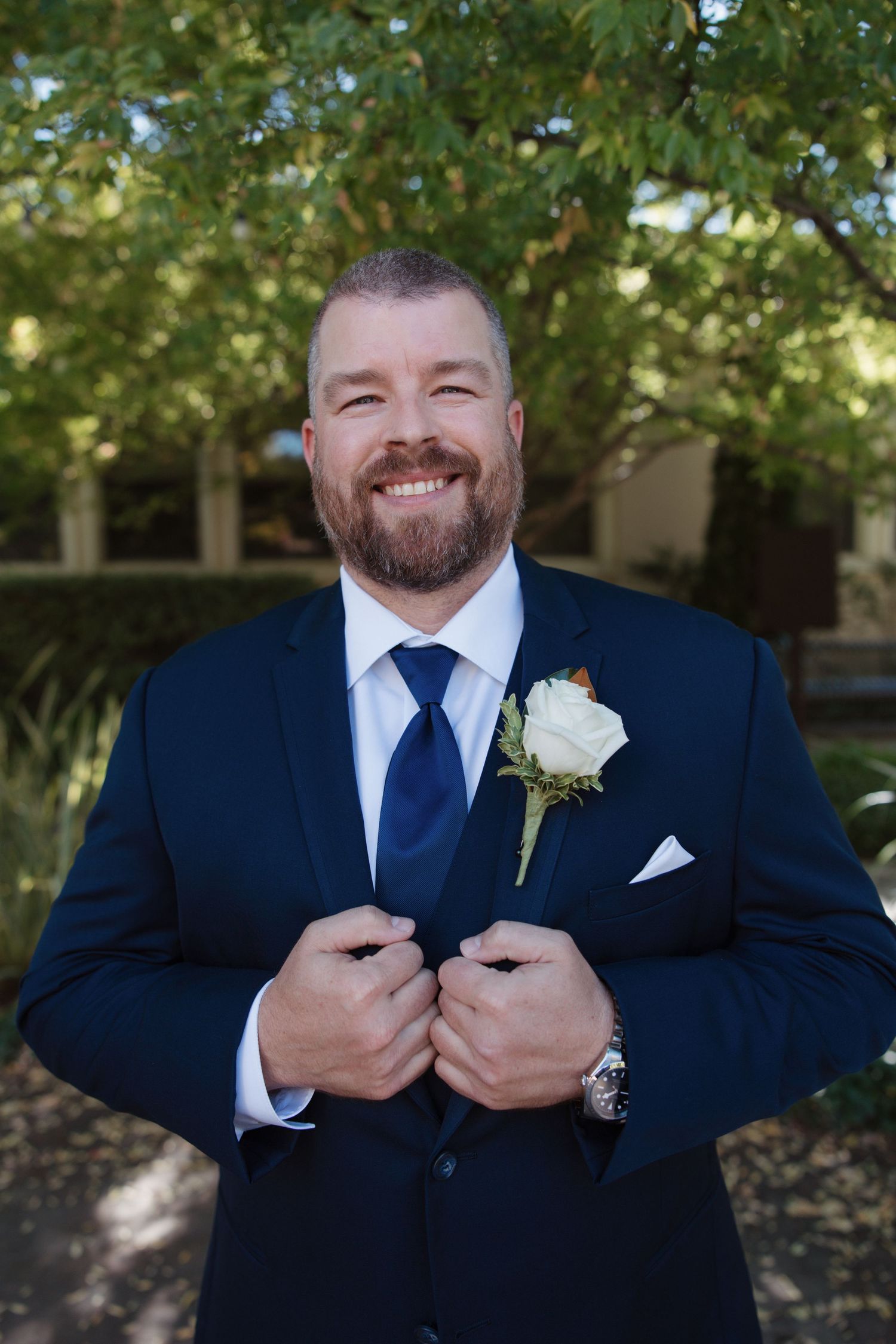 Smiling bearded groom in navy suit with white rose boutonniere adjusting his jacket outdoors.