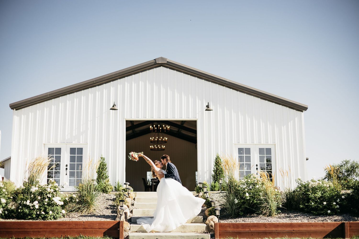 A bride and groom share a romantic dip kiss in front of a white barn wedding venue on a sunny day.