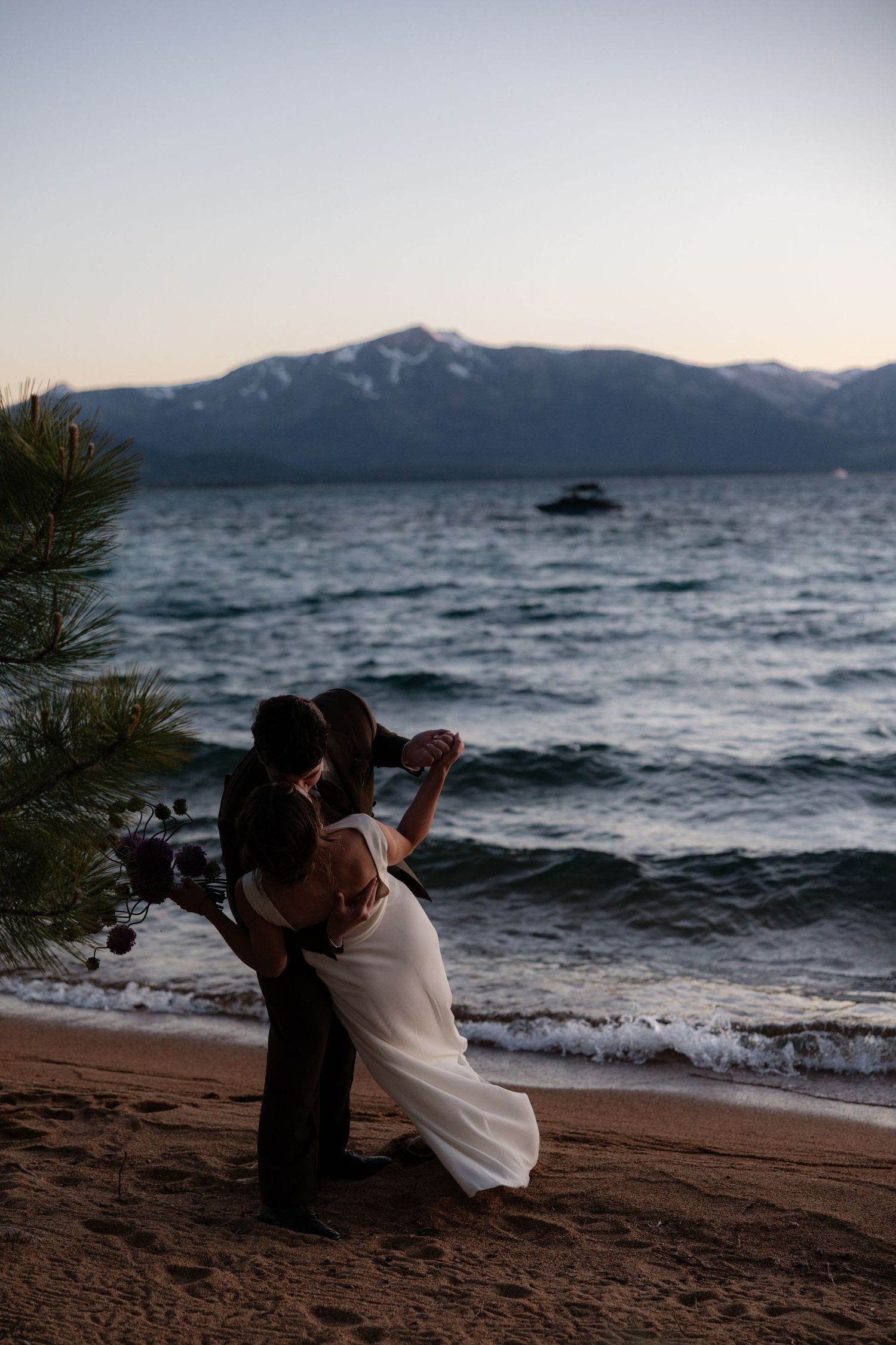 Bride and groom share a kiss on a sandy beach at dusk with snow-capped mountains in the background.