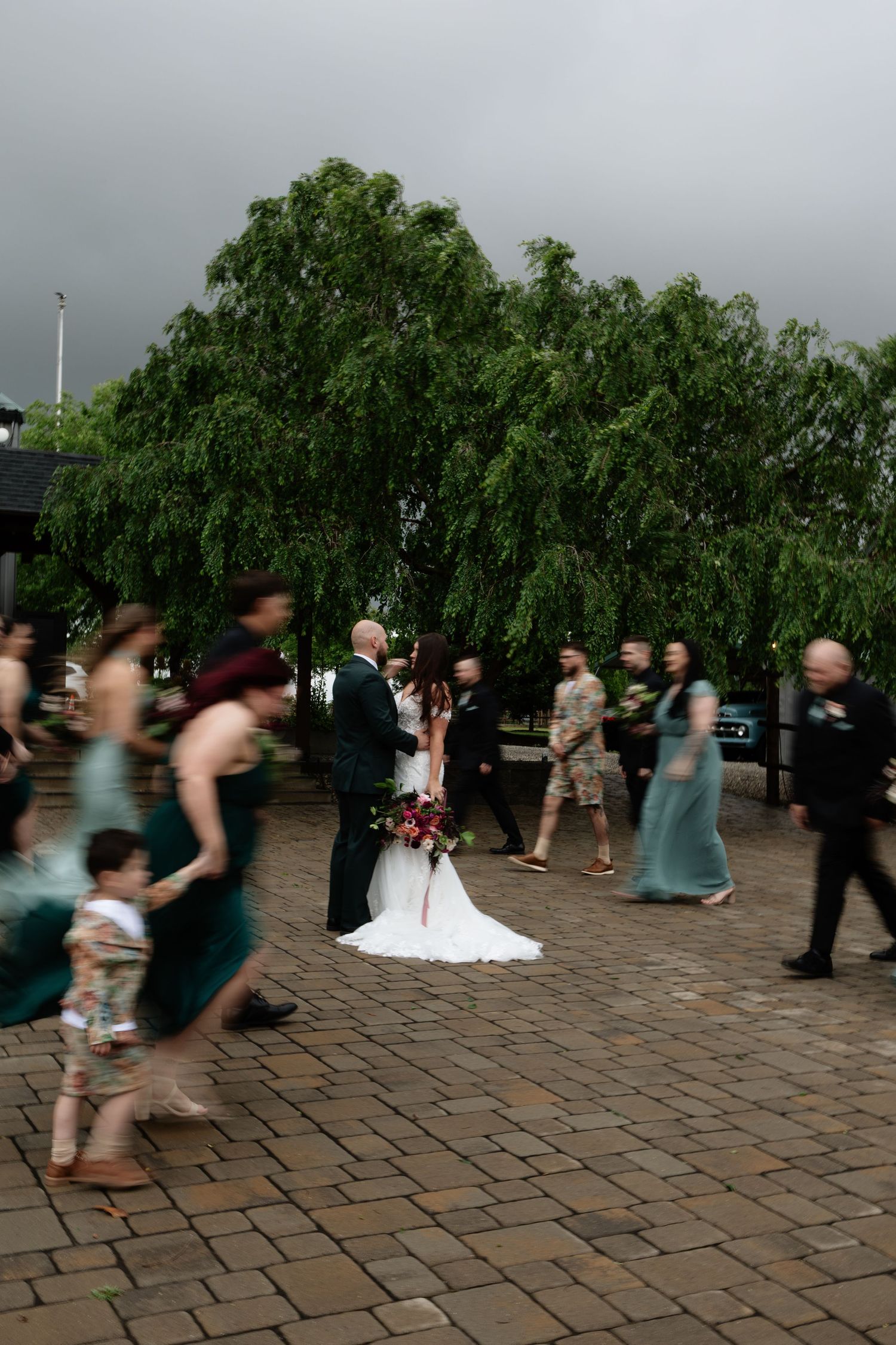 Wedding party moves across brick courtyard under stormy skies near a weeping willow tree.
