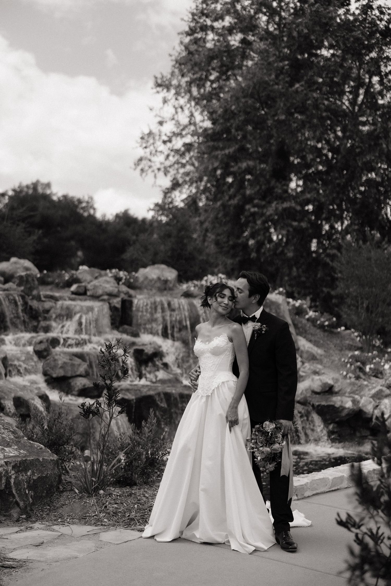 Bride and groom embrace near a romantic waterfall backdrop in a black and white wedding portrait.