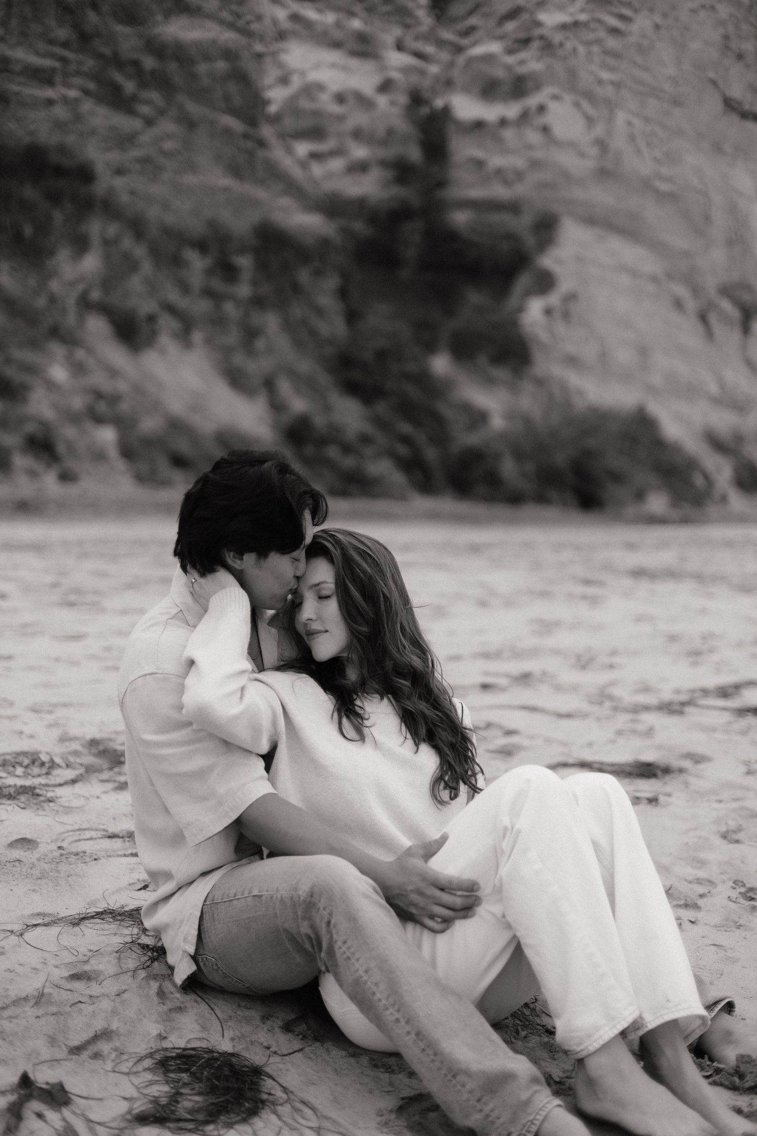 Black and white romantic photo of a couple sitting together on sand, leaning close against rocky cliffs.