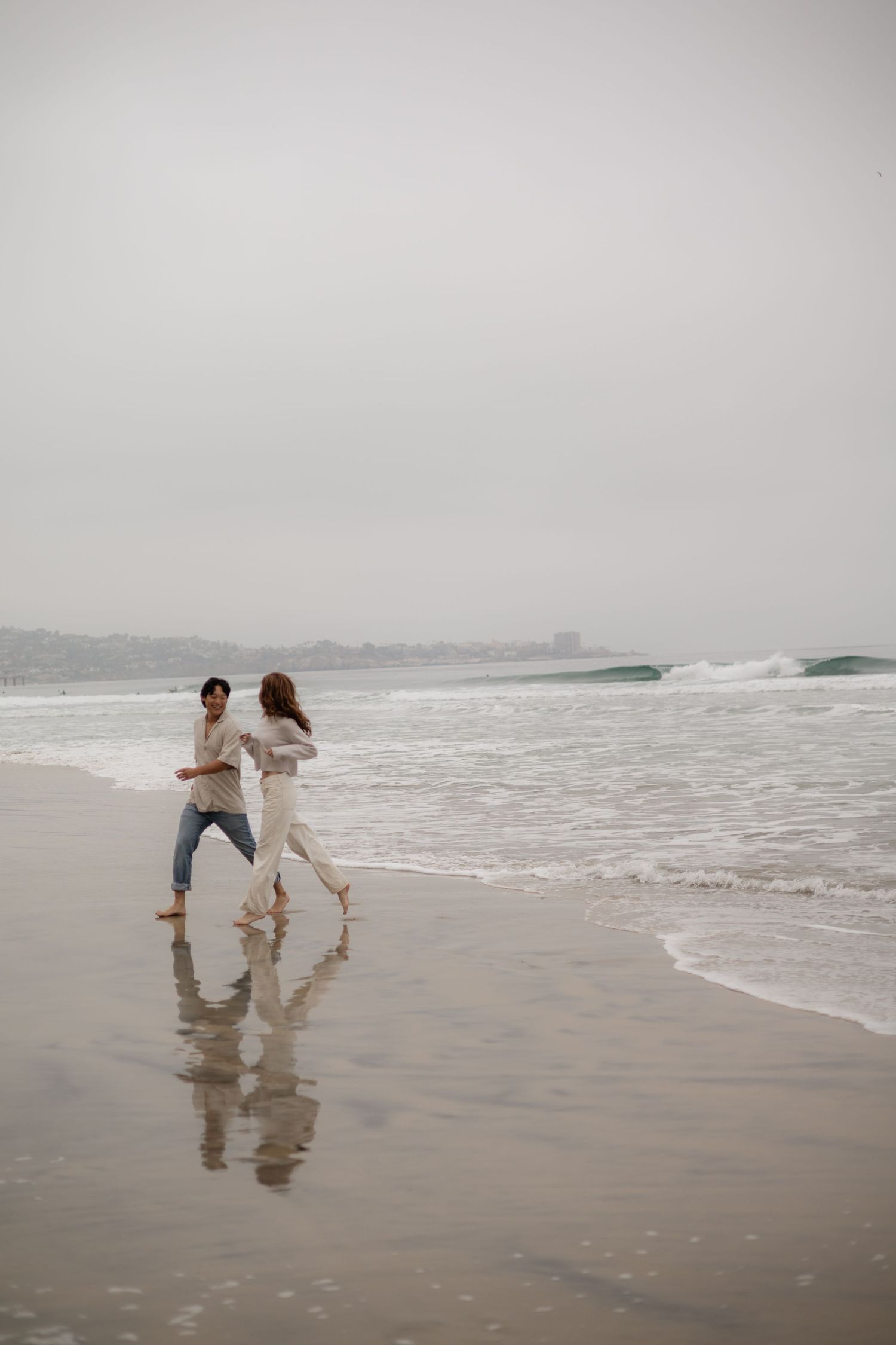 Couple running joyfully along a misty beach shoreline, waves crashing beside them on an overcast day.