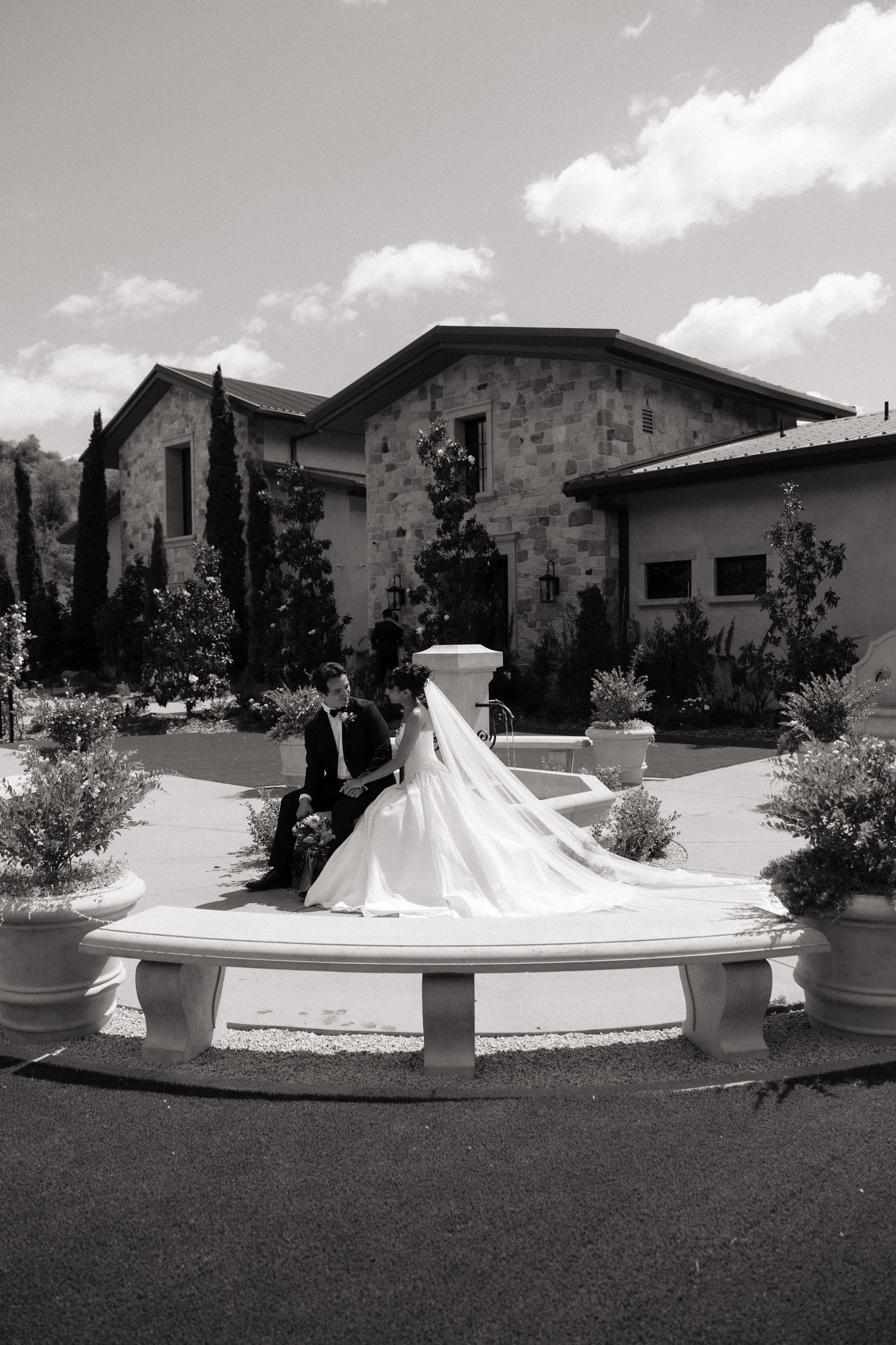 Bride and groom sit together on a white fountain outside a stone wedding venue in black and white.