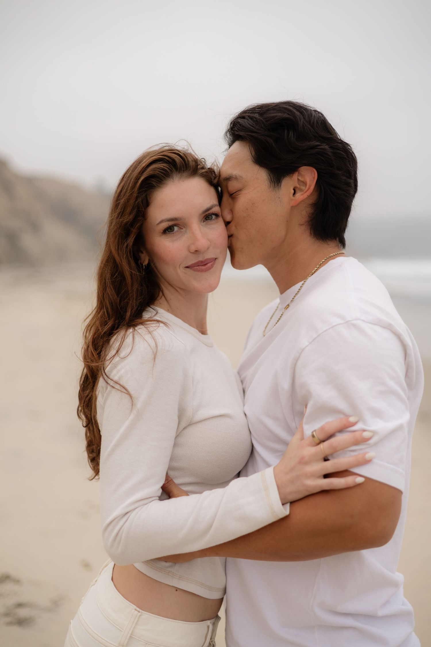 A couple in white outfits embracing on a misty beach, man kissing woman's cheek as she smiles at camera.