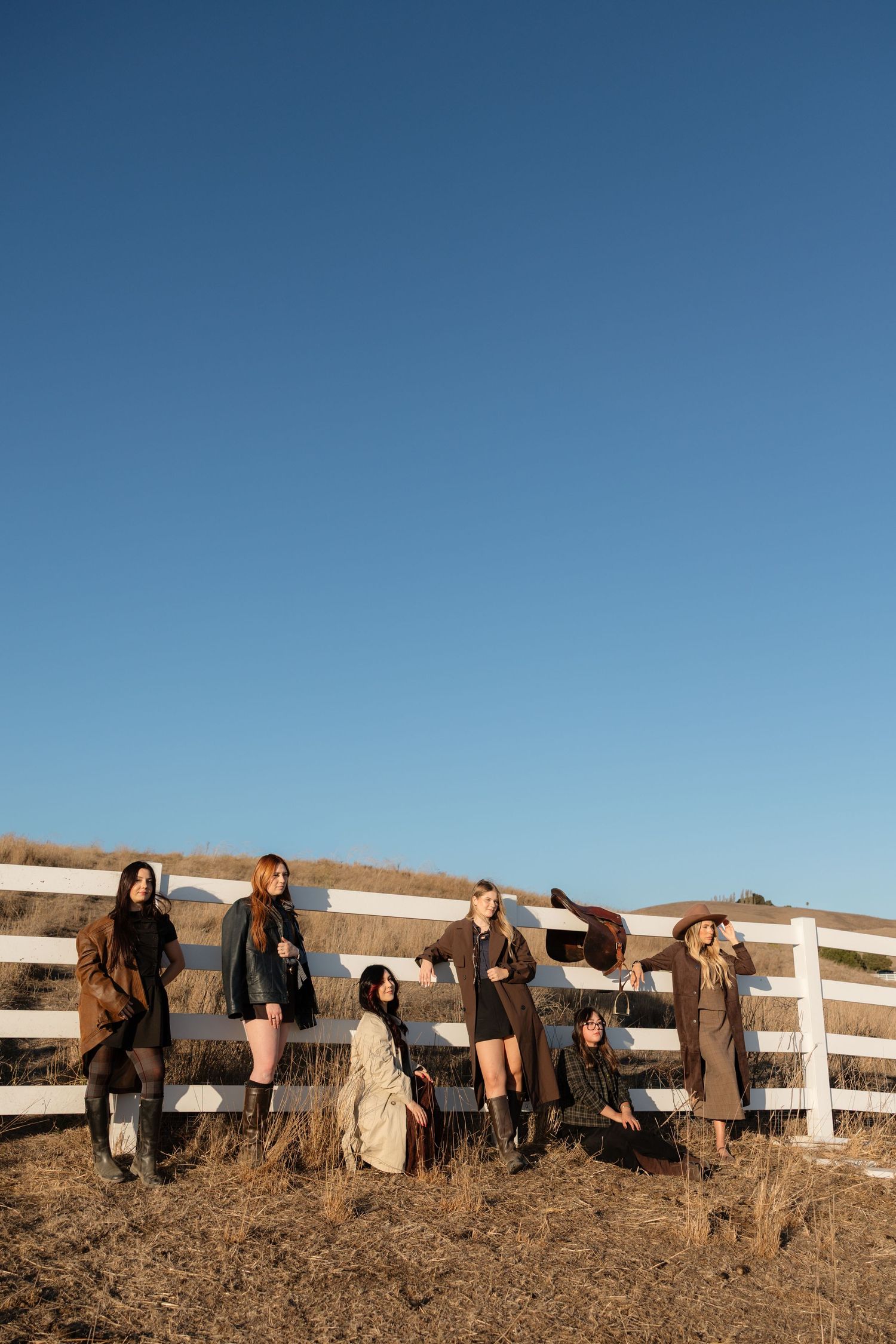 Six women in fall outfits pose against a white wooden fence under a clear blue sky on a rural hillside.