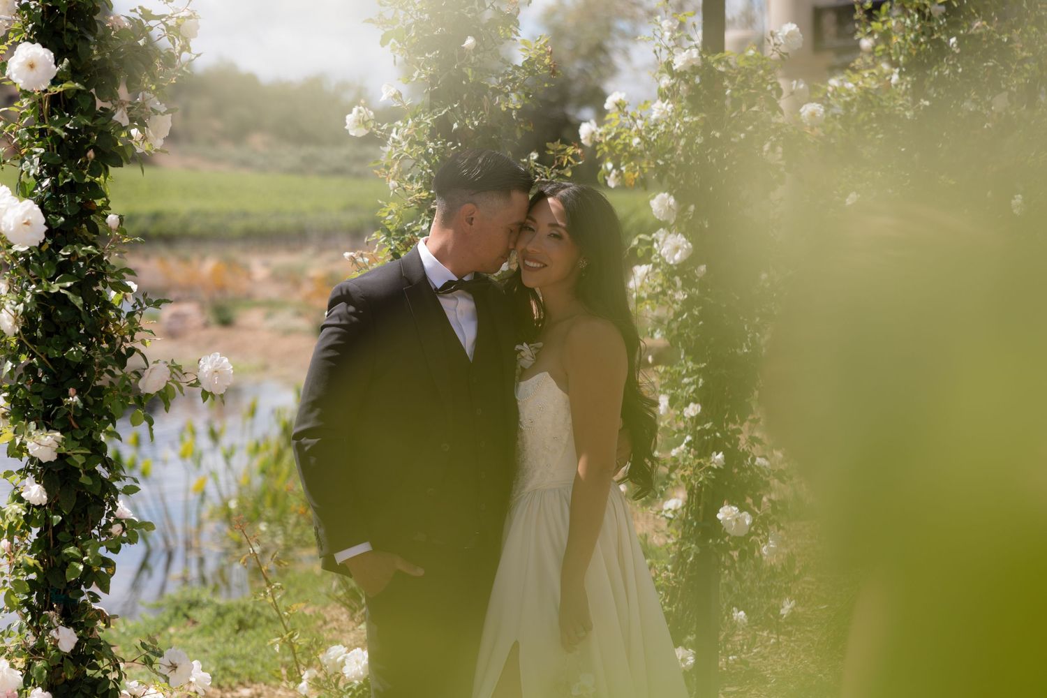 Romantic bride and groom kissing under white rose garden arch in soft sunlit wedding photo.
