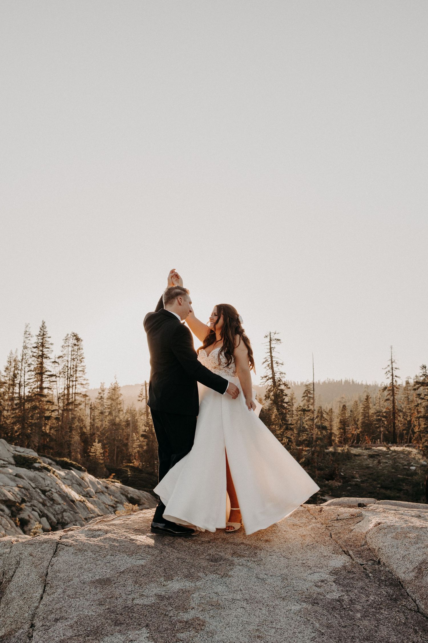 Bride and groom dancing on rocky mountain summit at golden hour surrounded by pine trees.