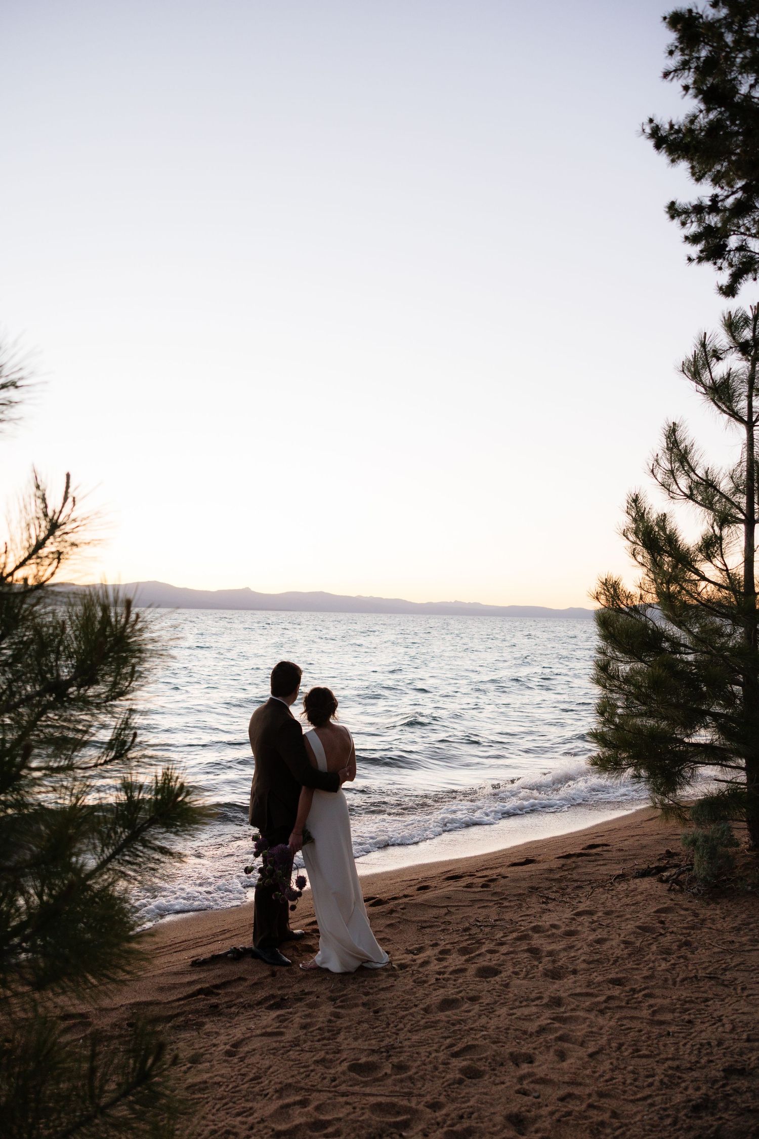 Couple in wedding attire standing on sandy beach at sunset, framed by pine trees with calm lake views.