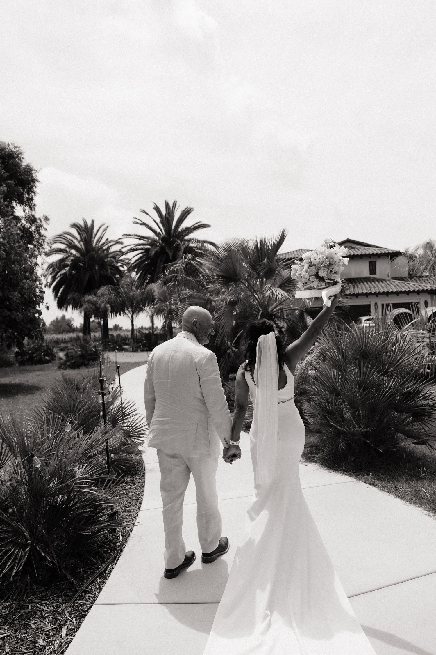 Black and white photo of bride and groom walking hand in hand past tropical palms toward a Spanish-style wedding venue.