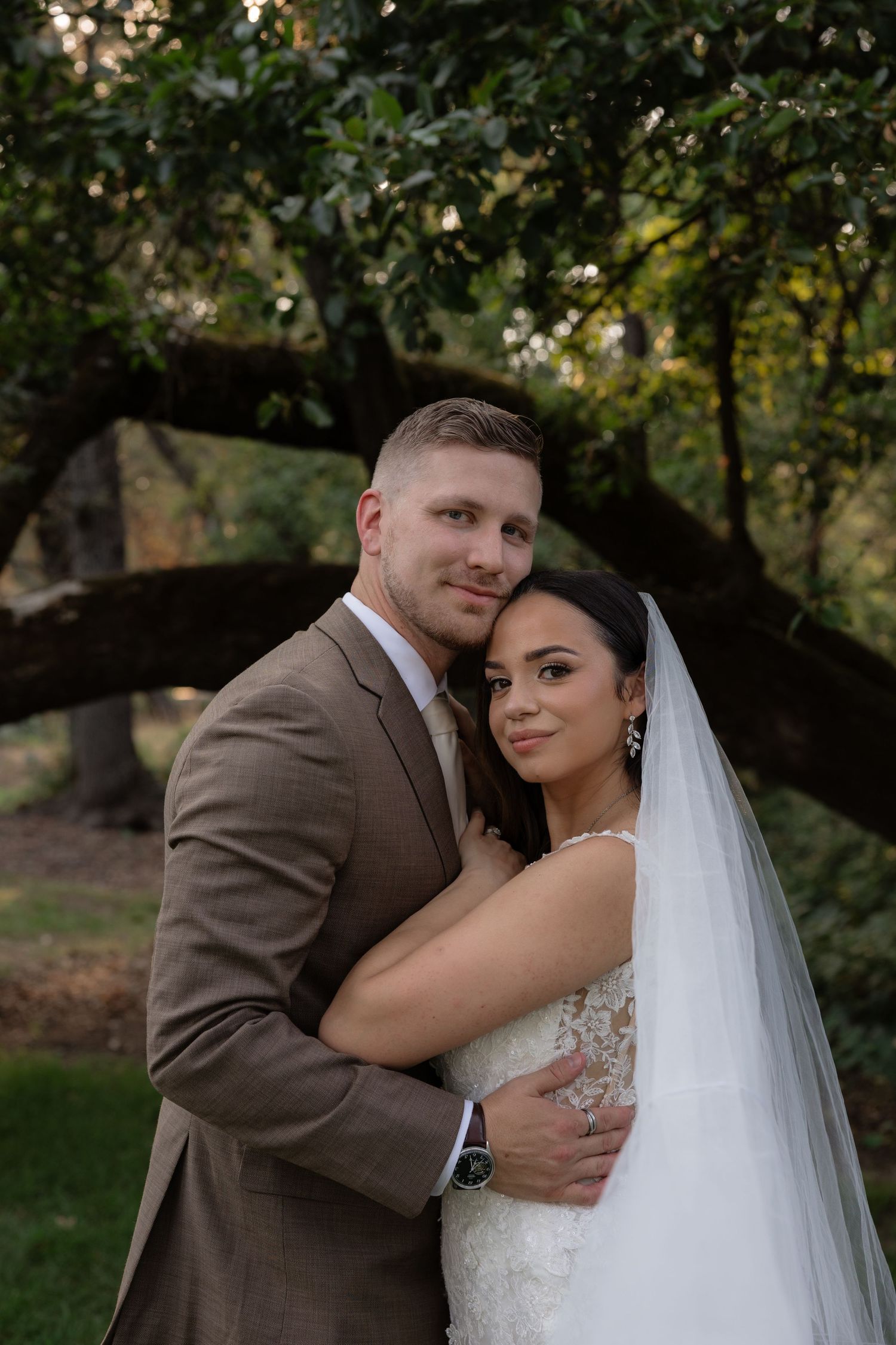 A bride and groom embrace outdoors under a large tree, wearing a white gown with veil and tan suit.