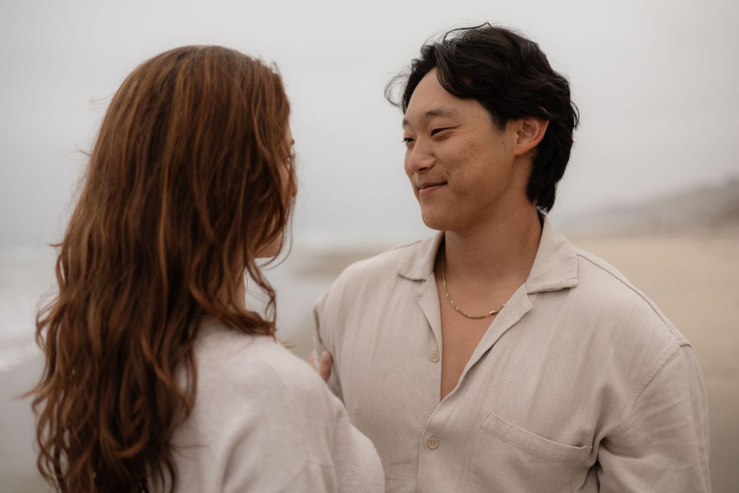 Couple sharing a romantic moment on a misty beach, smiling at each other in matching beige linen shirts.