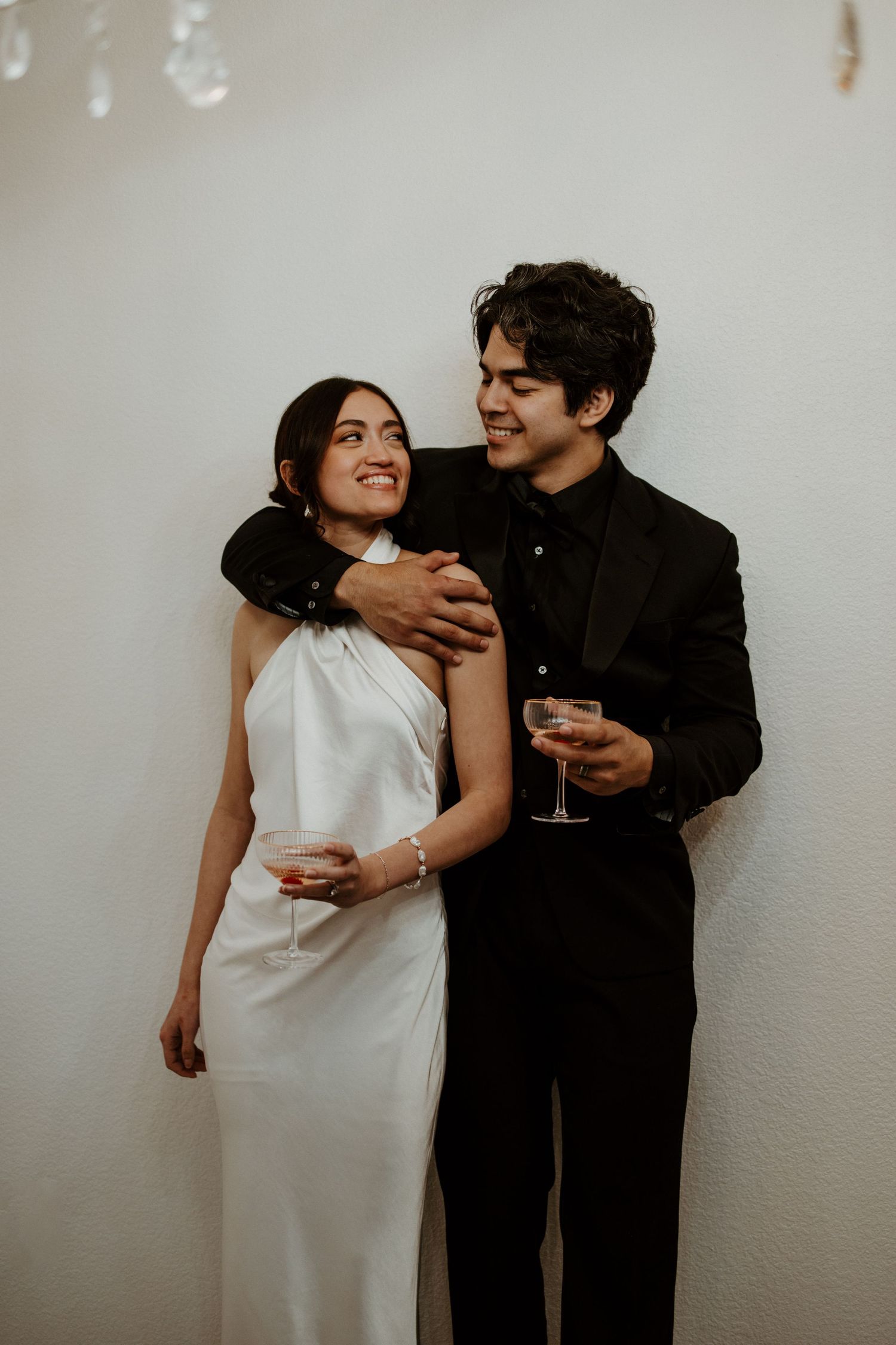 Smiling couple in wedding attire holding champagne glasses against a white wall.