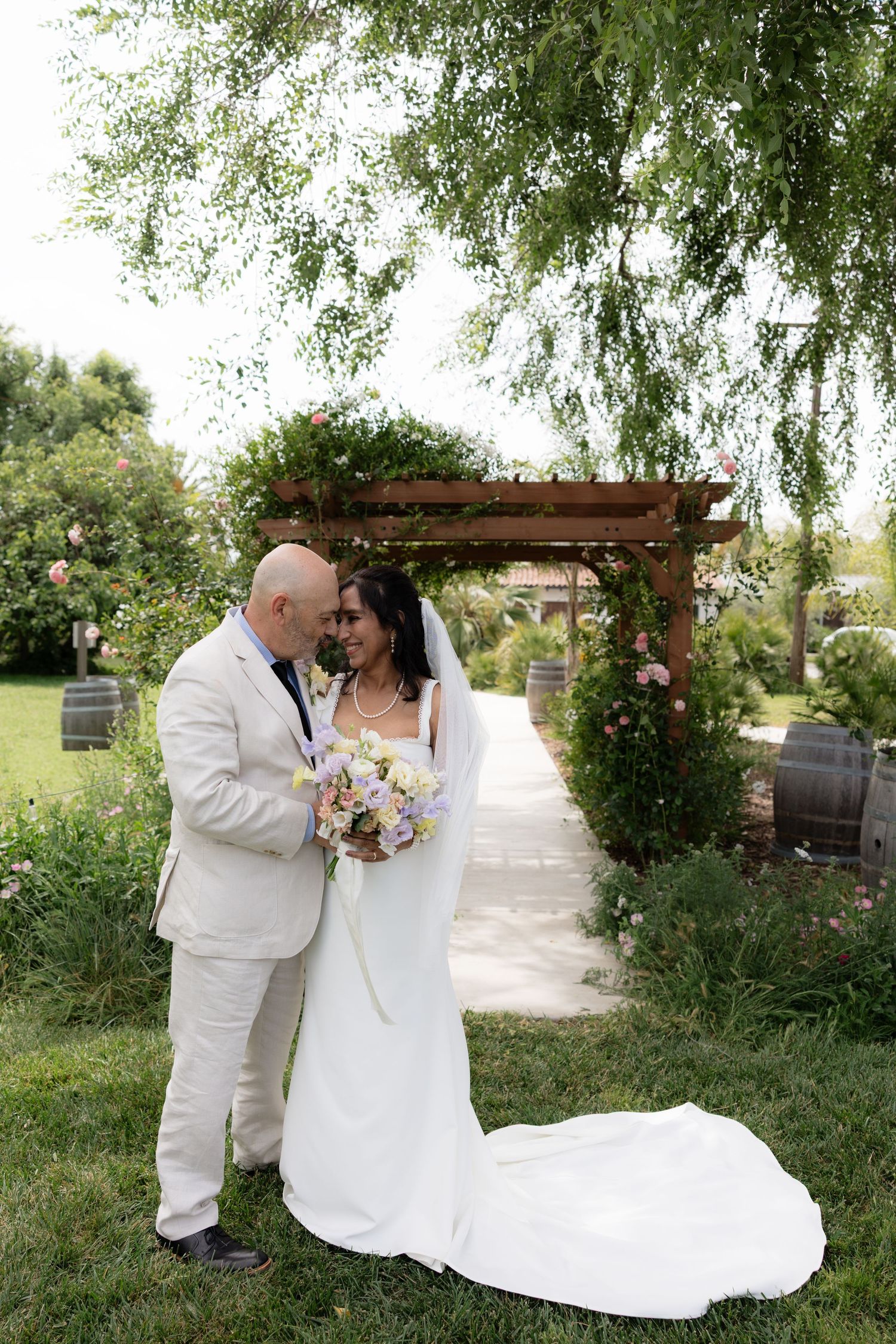 Bride and groom share a romantic moment under a wooden pergola surrounded by lush garden greenery.