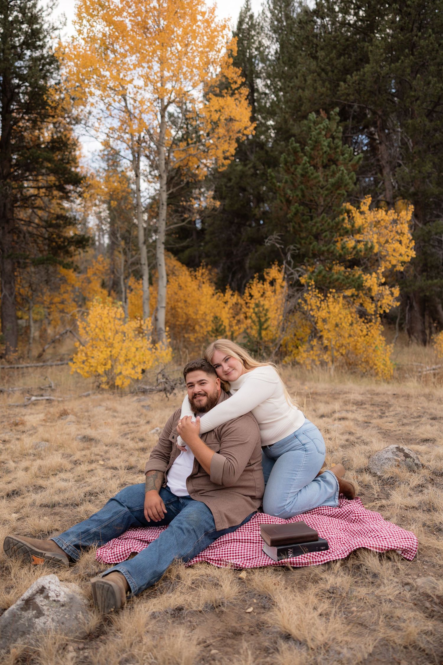 Couple sitting on a red picnic blanket in a fall forest with golden aspen trees, sharing a warm embrace outdoors.