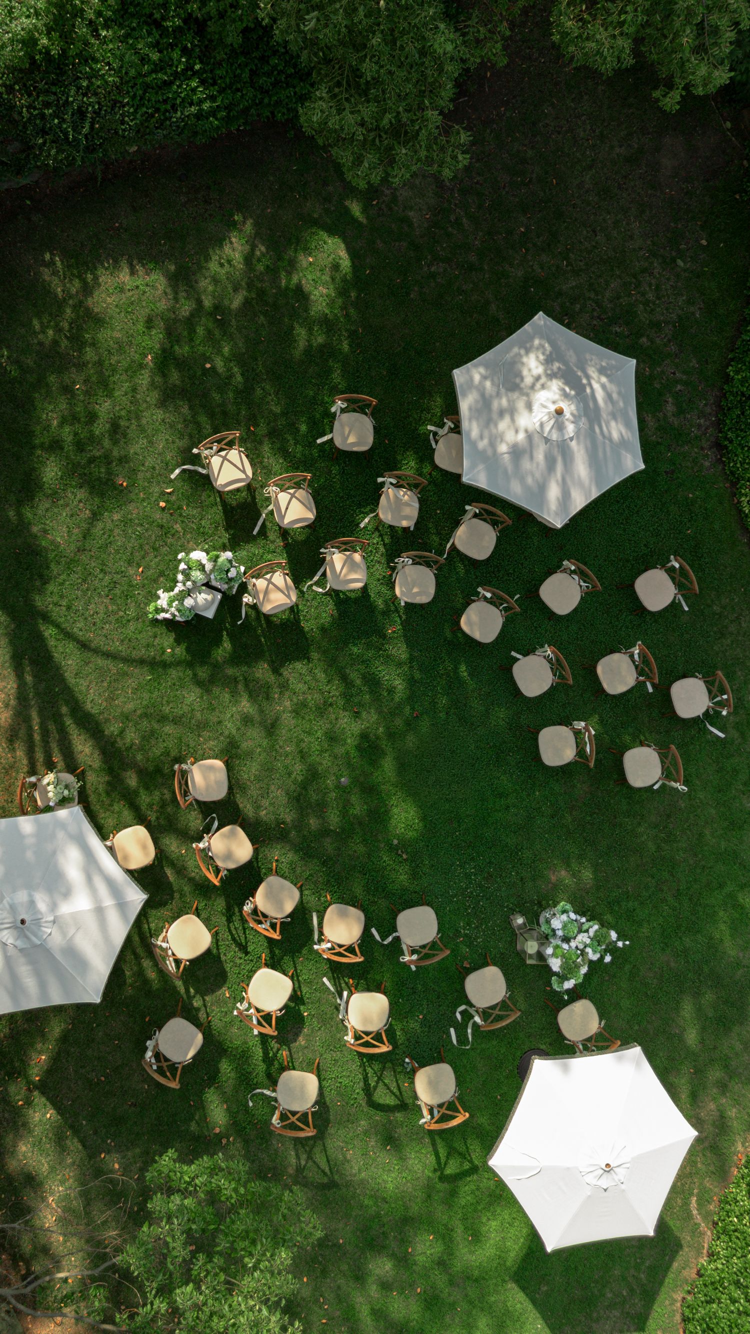 Aerial view of an outdoor wedding ceremony setup with wooden chairs arranged on green grass, flanked by white patio umbrellas.