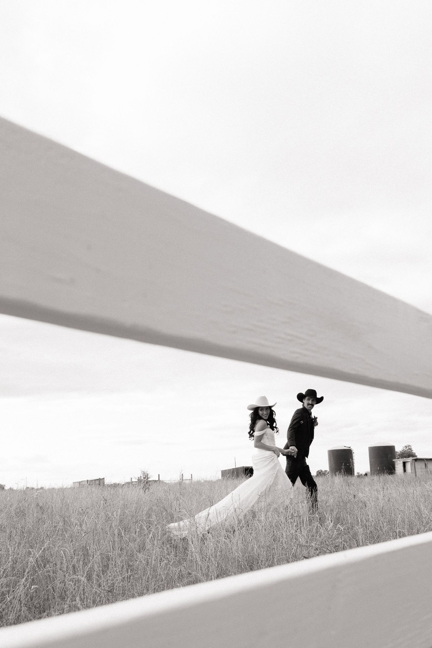 Black and white photo of wedding couple in cowboy hats walking through a field viewed through a fence.