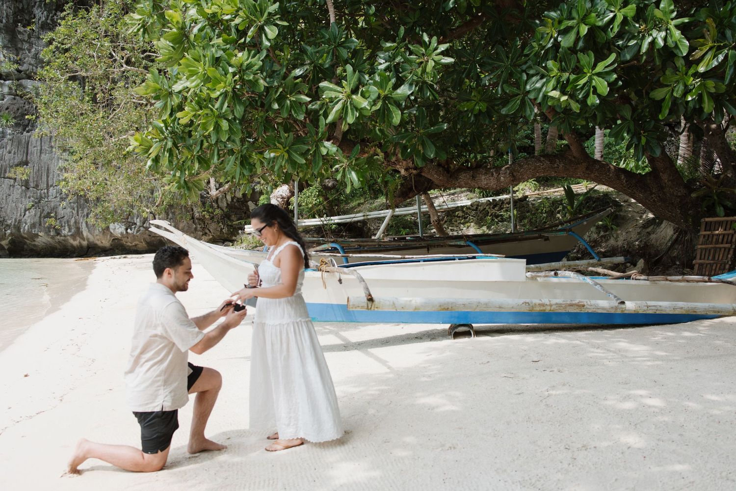 Romantic beach proposal scene with man kneeling before woman in white dress near a traditional boat.