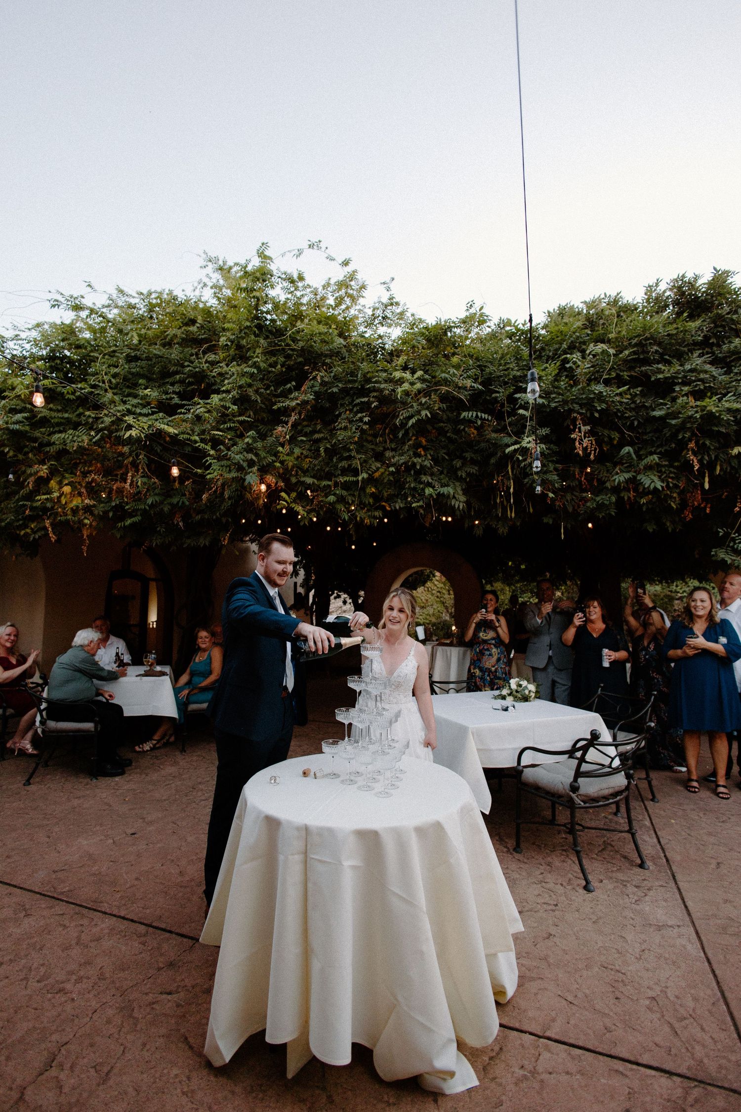 Bride and groom pour champagne into a tower of glasses at their outdoor evening wedding reception.