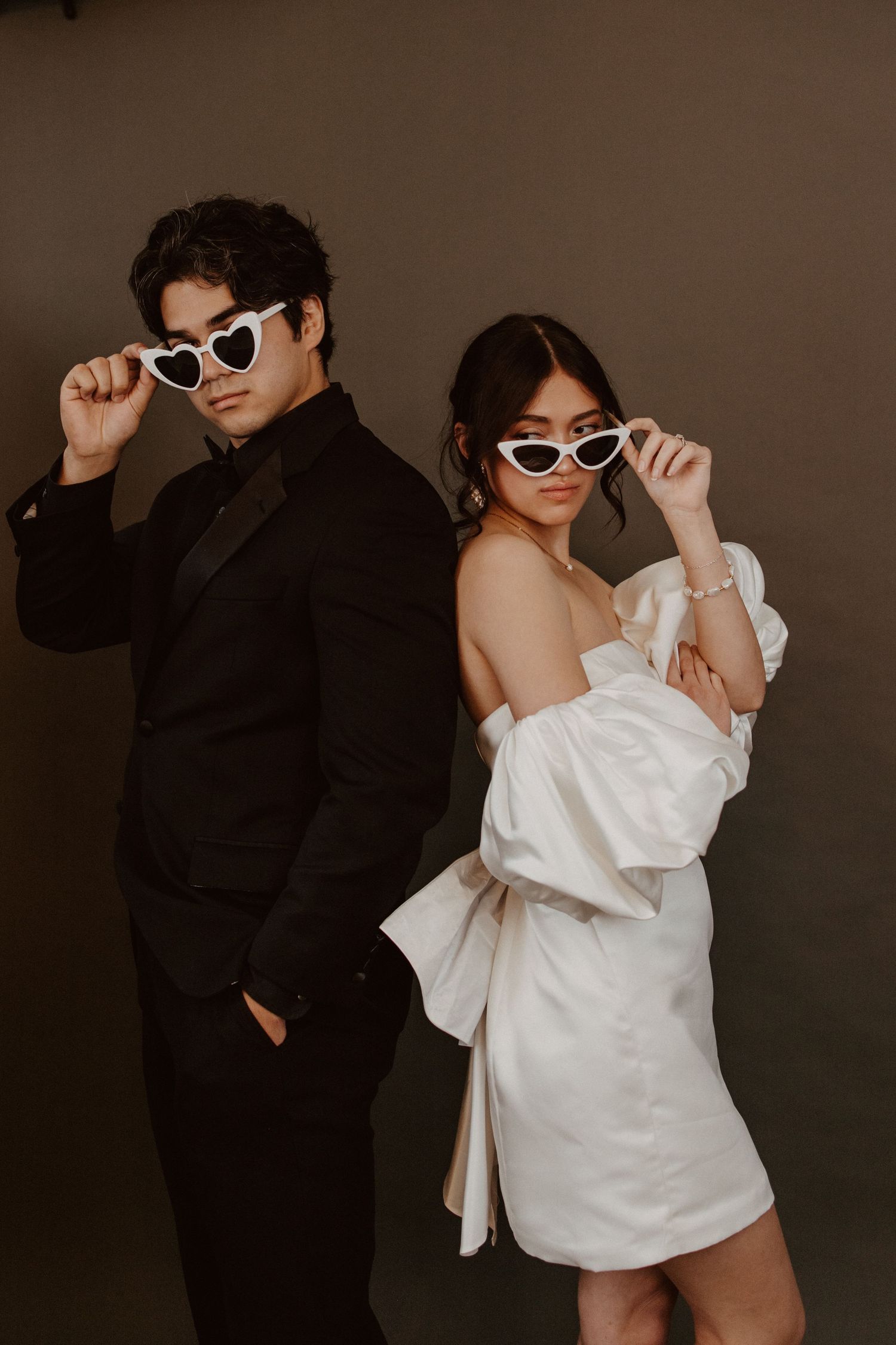 Stylish couple in black suit and white mini dress posing with heart-shaped sunglasses.